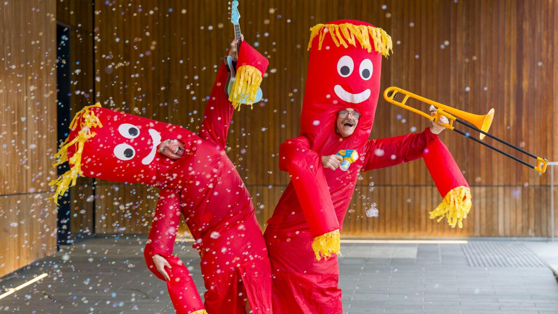 Two people in red tube costumes with yellow fringing. One holds a violin and the other holds a trombone. Confetti erupts before them and one strikes a jaunty angle.