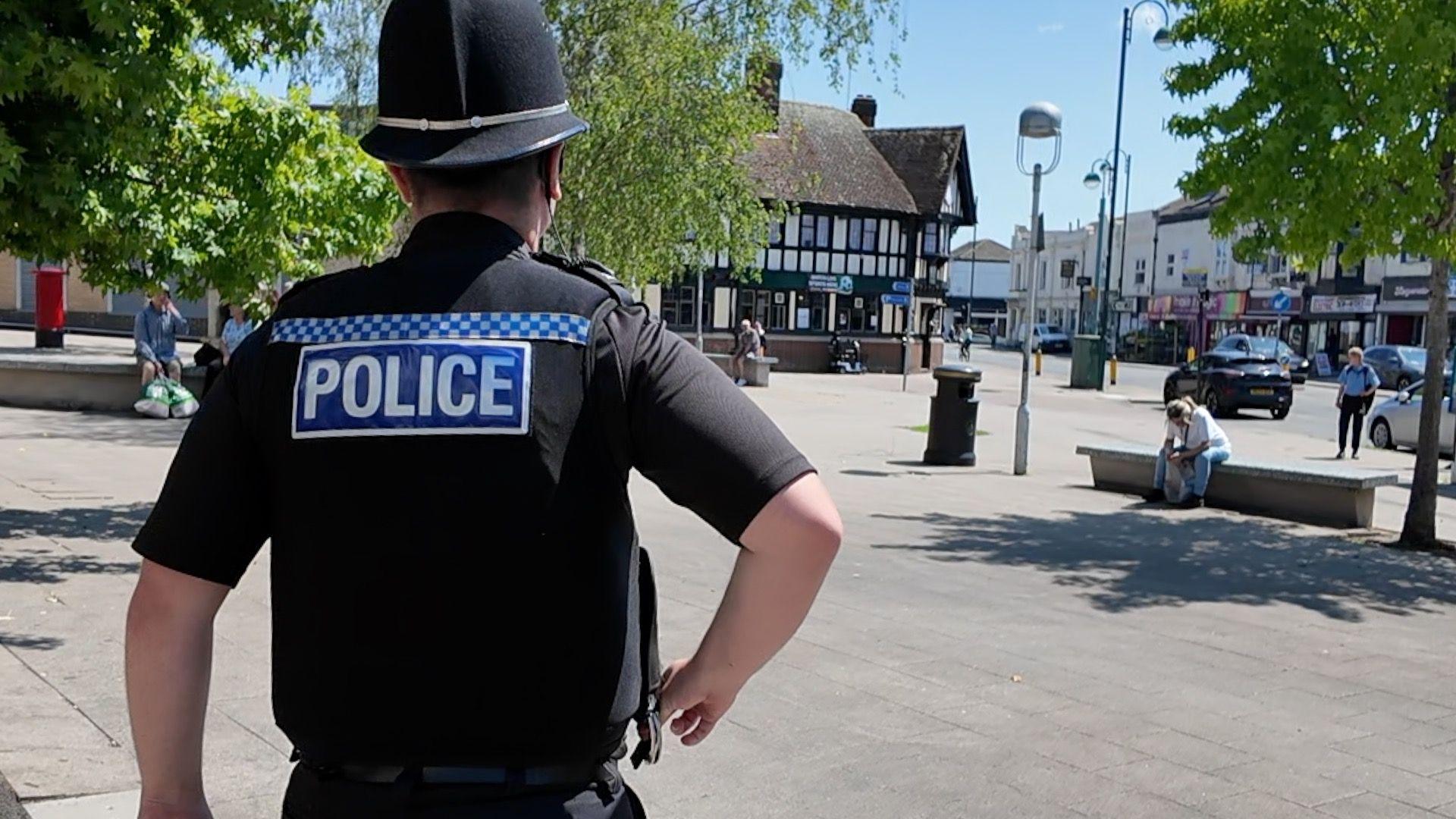The back of PC Byrne as he walks down Shirley high street on a sunny day. He is wearing black with Police written across his back in white letters. He is walking across a paved square surrounded by shop. There are people sitting on benches and three trees. 