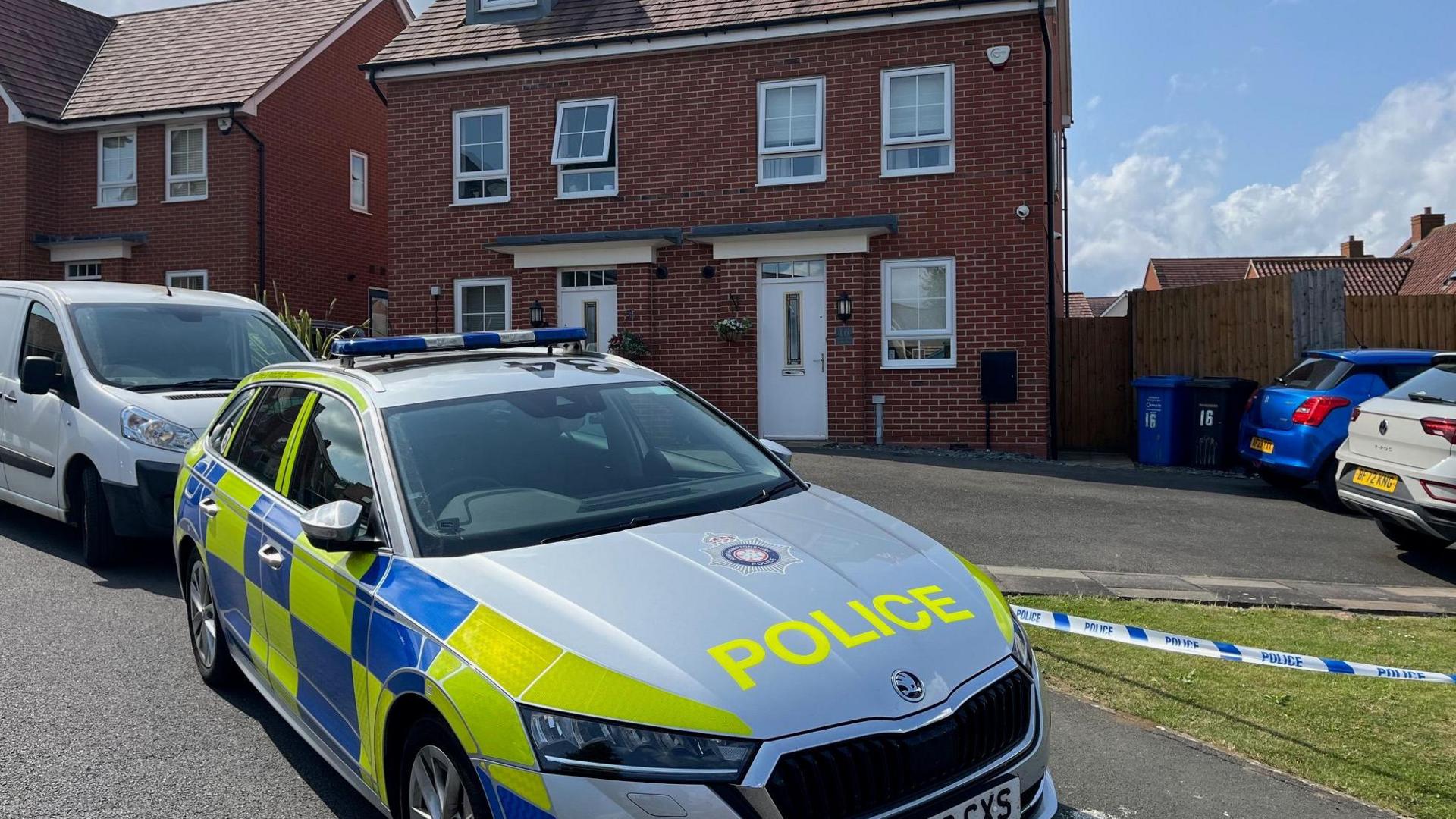 A police car outside a three-storey new build semi-detached house with white windows and door. There is police tape outside the Tarmac'd area in front of the house. Two cars are parked on this.