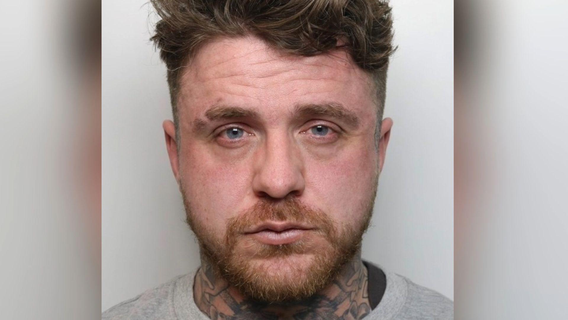 A police mugshot of Shane Davis. He is standing against a light-coloured background. He has brown hair and a beard, with tattoos around his neck.