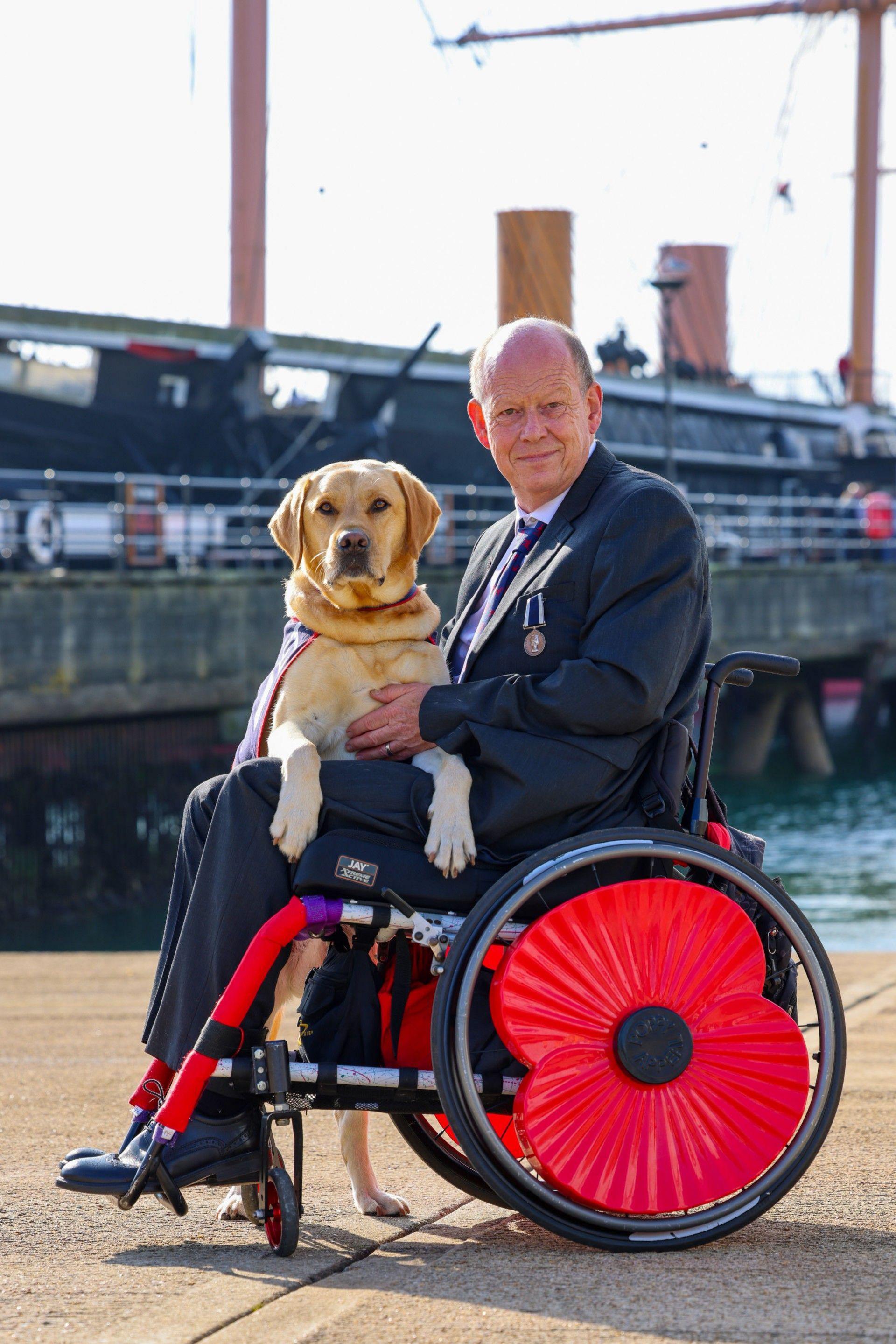 Allen Parton is in his wheelchair, which has large red poppies covering the wheel. He wears a black suit with a white shirt and navy blue and red printed tie and has a medal on his left breast pocket. His service dog, a golden labrador called Endal, is on his hind legs leaning over Allen's legs. HMS Warrior and the sea can been seen in the background.