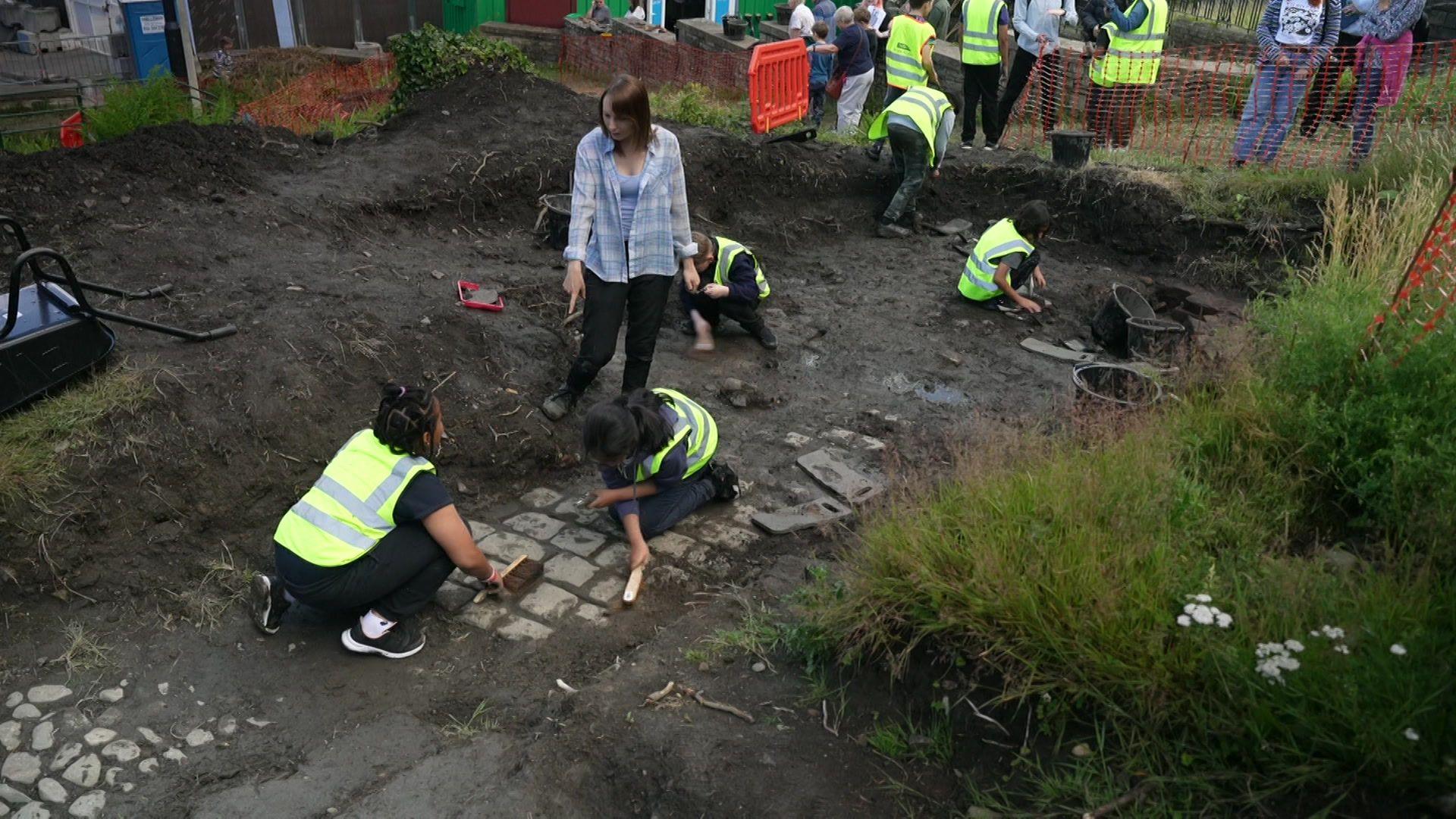 Image shows people in high-visibility tabards digging into the earth, uncovering what look like sections of cobbled streets below the soil