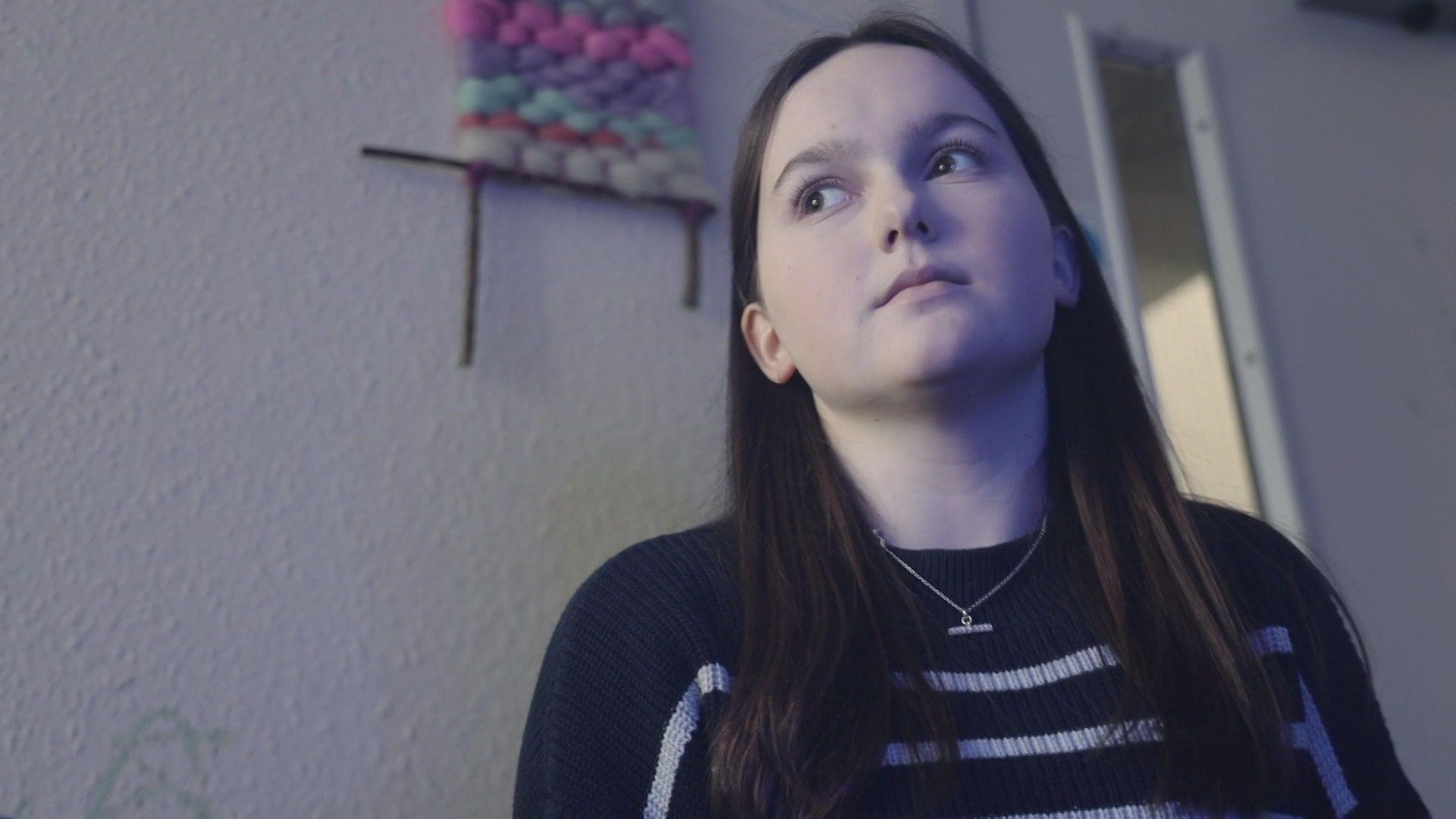 Megan mewn golygfa o'r ffilm Tyfu Mewn Galar - merch ifanc gyda gwallt hir tywyll mewn ystafell yn edrych tua'r pellter canolig