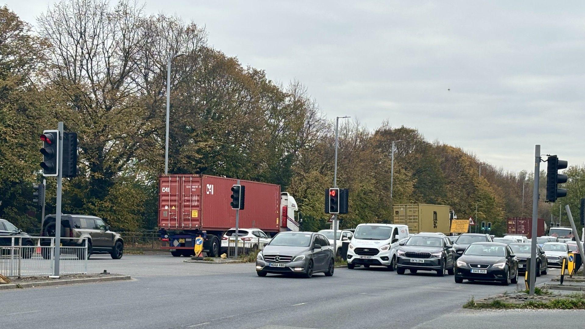 Lorries and cars travel through a busy dual carriageway junction. Trees line the road.