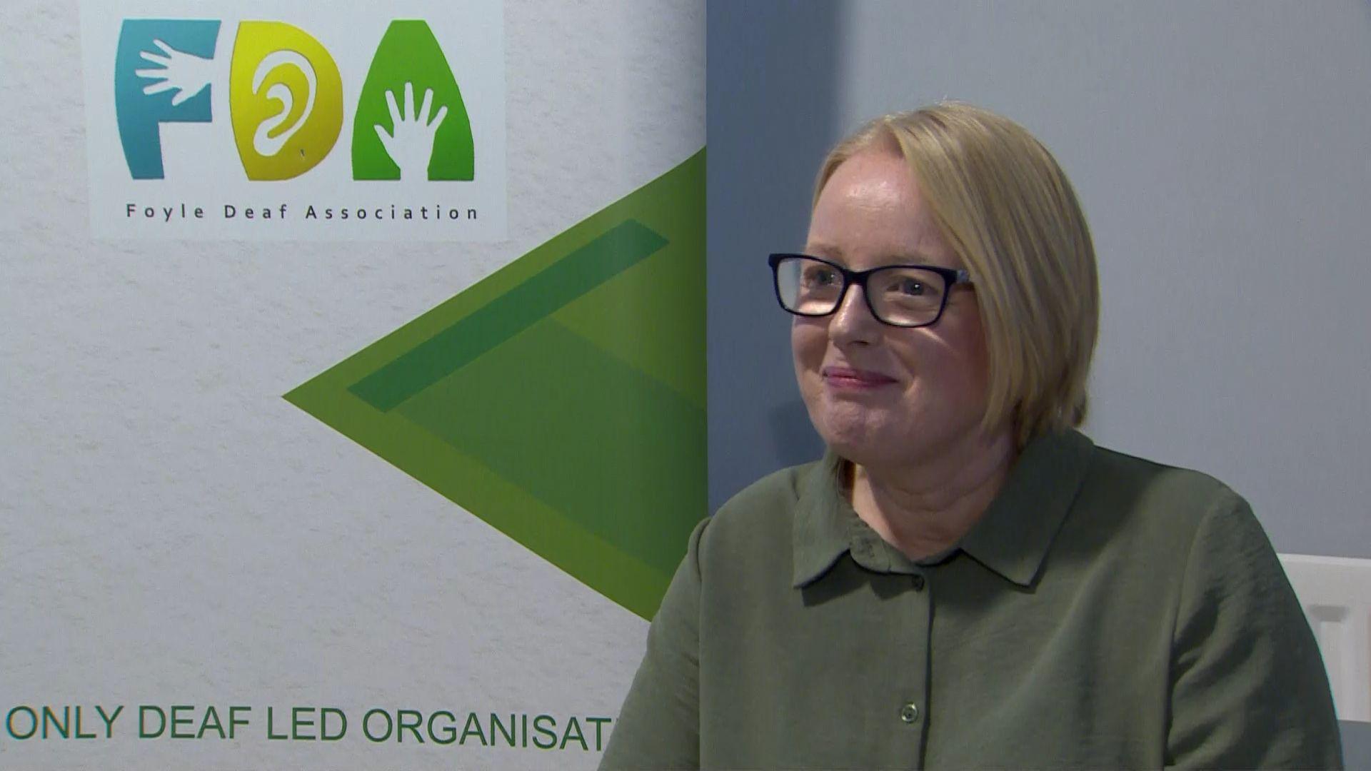 A woman with short blonde hair and glasses sits smiling looking to the left of the camera. She is wearing a green top and in the background is a standing poster board for Foyle Deaf Association 