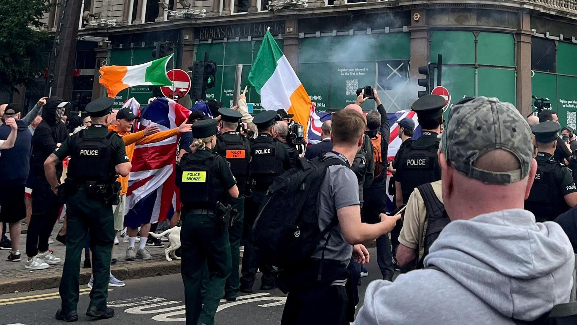 Police and protesters standing close to each other. Some protesters are holding union flags and some are holding Irish tri-colour flags