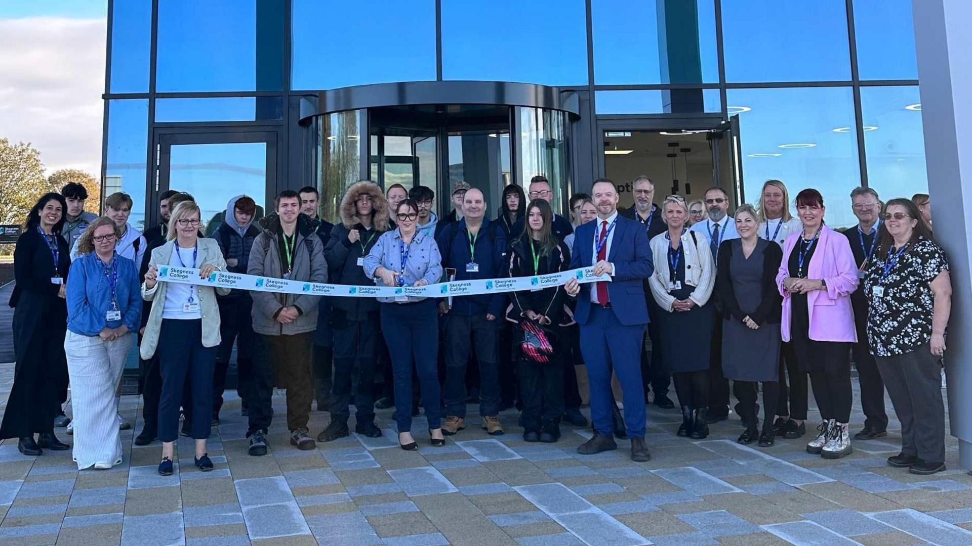 A group of people stood outside a glass building. They are cutting a ribbon.
