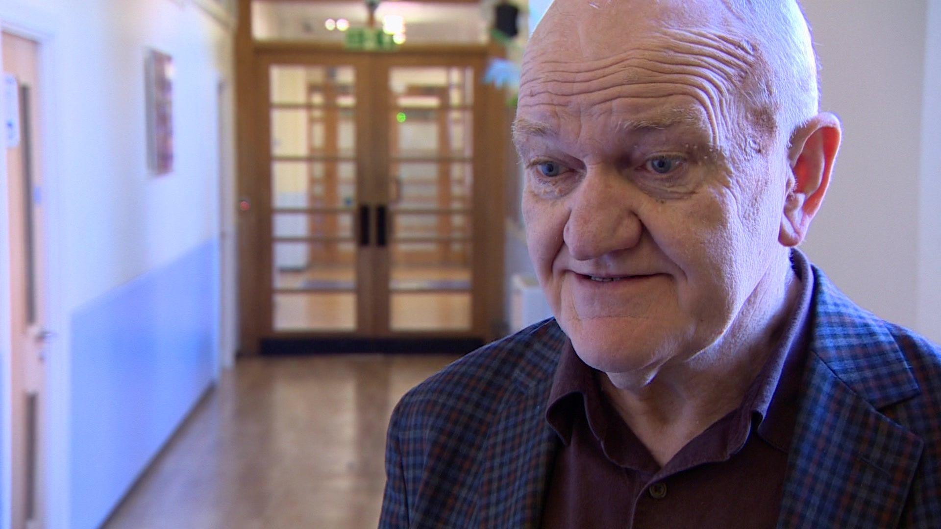 Elderly man standing in a school corridor. He's wearing a dark blue, close checked jacket and a maroon colour shirt underneath