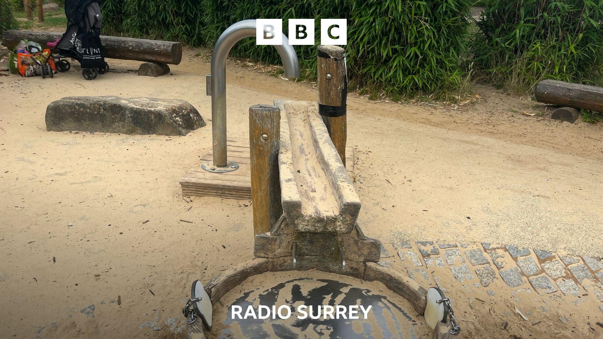 A wooden fountain play structure set within a sand pit. At the top of the structure is a large metal pip designed to poor water, but no water is coming out.