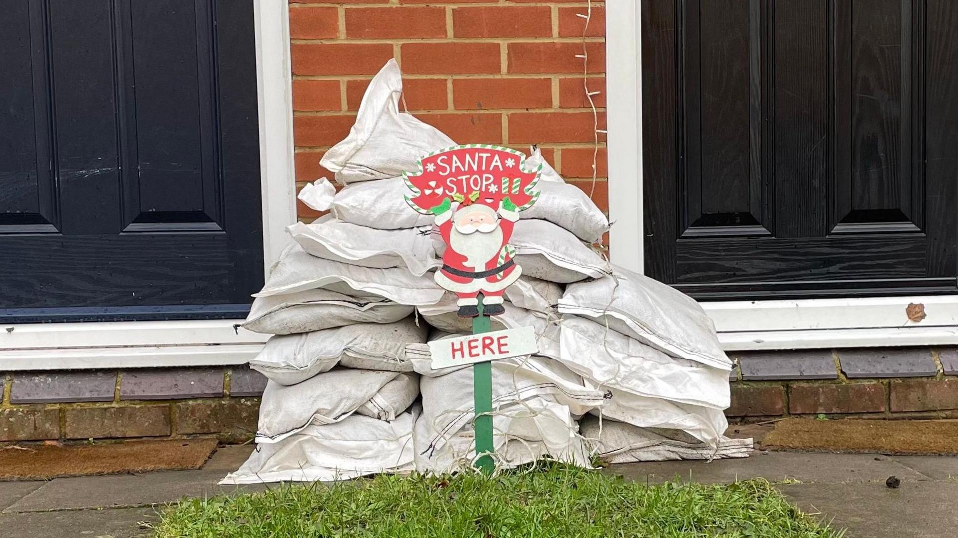 A 'Santa stop here' sign in front of a pile of sandbags. There is a small patch of grass visible in front of the sign. Behind the sandbags the bottom half of two front doors can be seen.