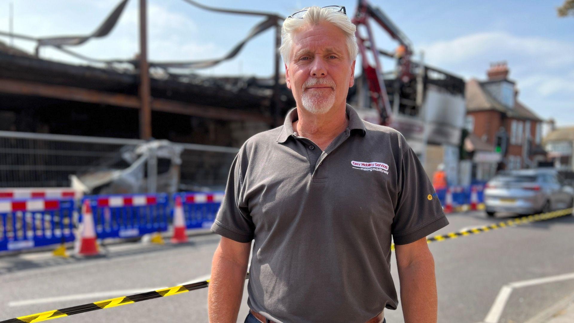 A man looking straight at the camera while wearing a grey polo top with the words "Easy Mobility Services" on the chest. He is standing in front of a building which has been destroyed by a fire.
