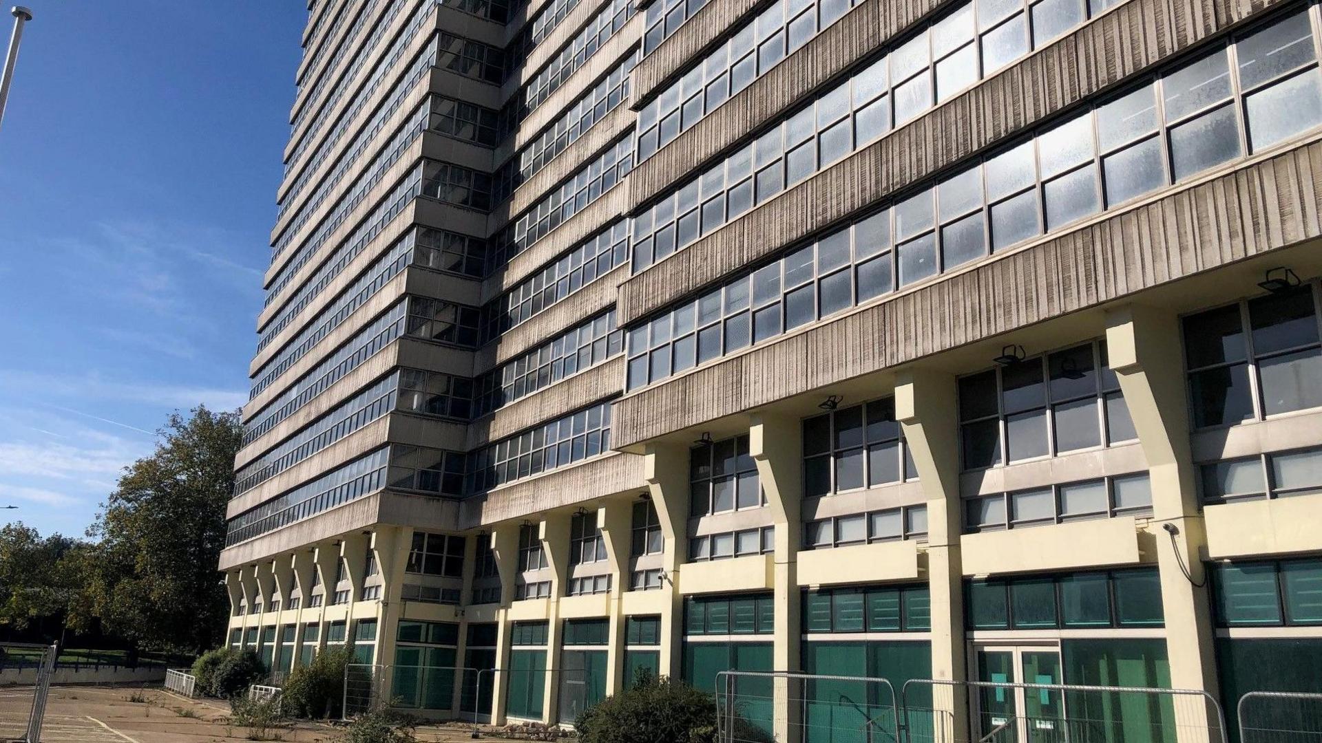 Alexander House in Southend. It is an empty, concrete office block, with rows of windows on each floor. It is too tall to fit into the picture. At the front of the building, there are empty parking spaces with overgrown weeds.