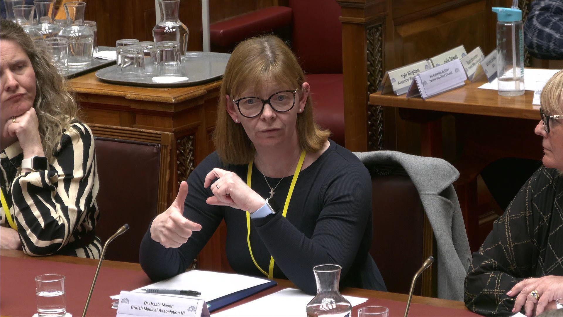 A red haired woman with a black long sleeve top, wears a yellow lanyard. She sits at a red leather desk with her elbows on the table. Her grey jacket sits on the red chair she is sitting on. She wears glasses and silver earings