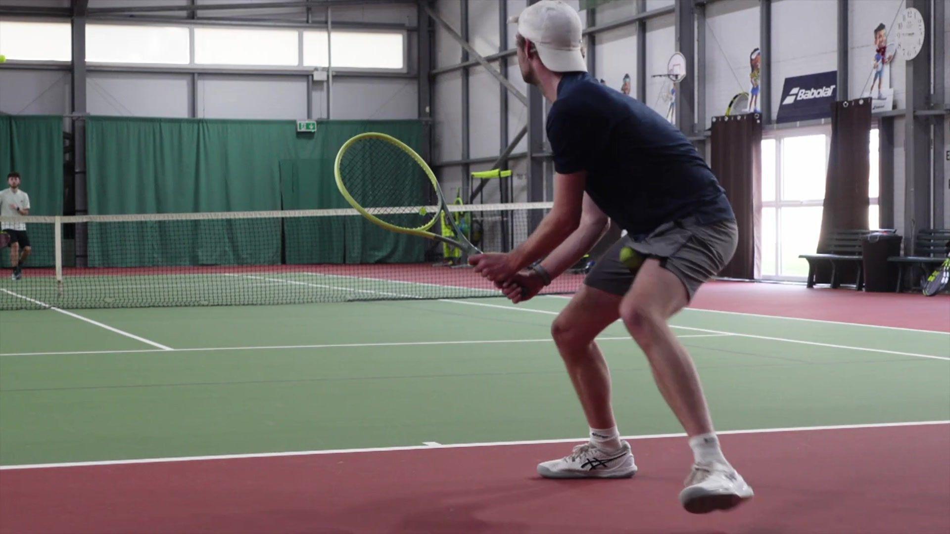 A man with a backwards white baseball cap plays tennis at an indoor court. 