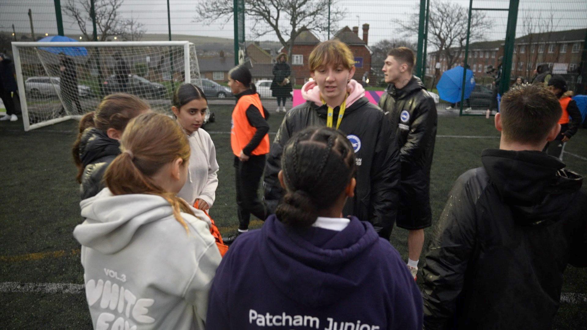 Jaime Quinn chatting to a group of girls who she coaches standing on an astro turf football pitch. There's a goal behind them and metal fence