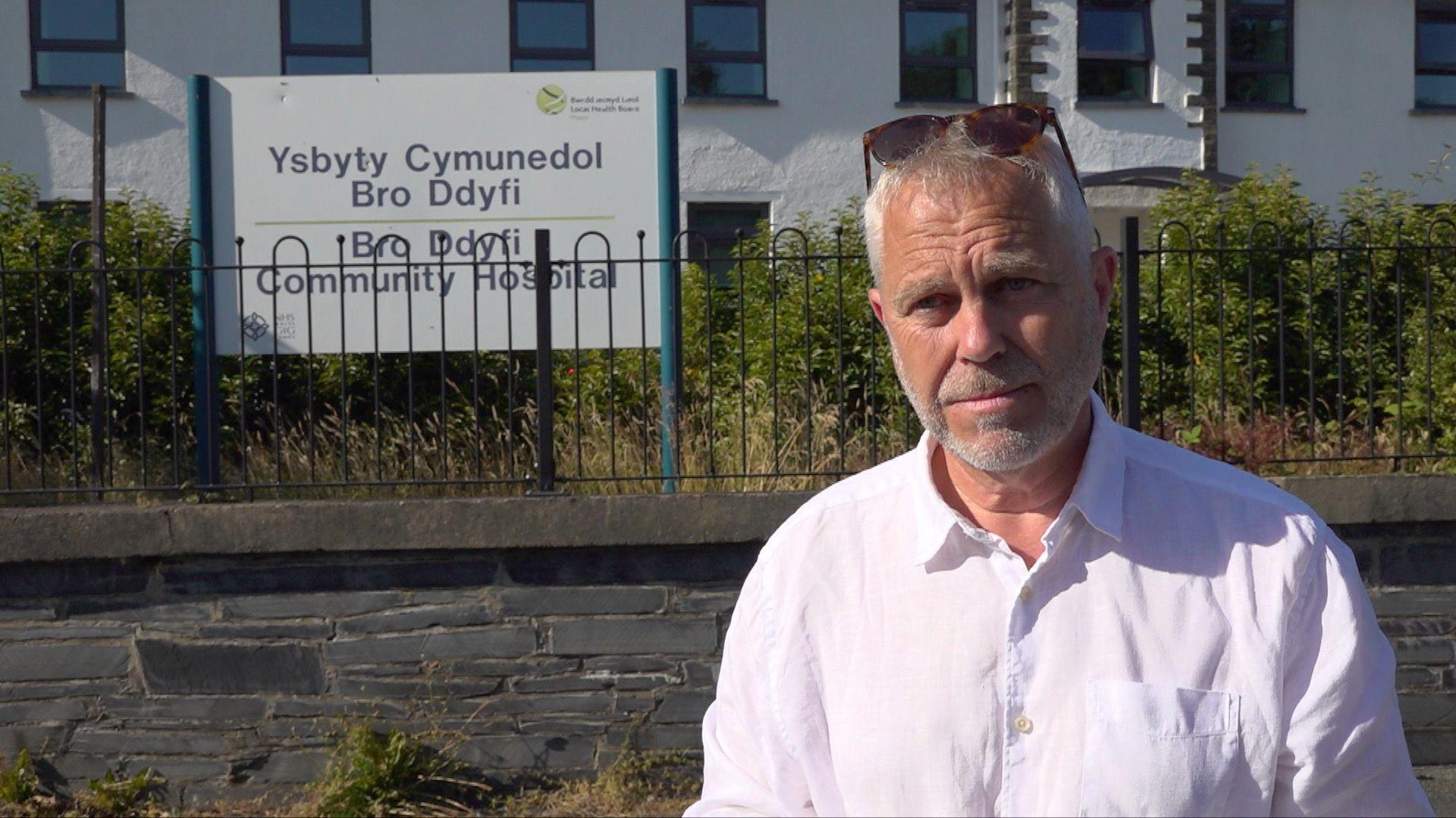 Yr aelod seneddol Elwyn Vaughan yn sefyll o flaen ysbyty cymunedol Bro Ddyfi yn gwisgo crys gwyn a sbectol haul ar ei ben.