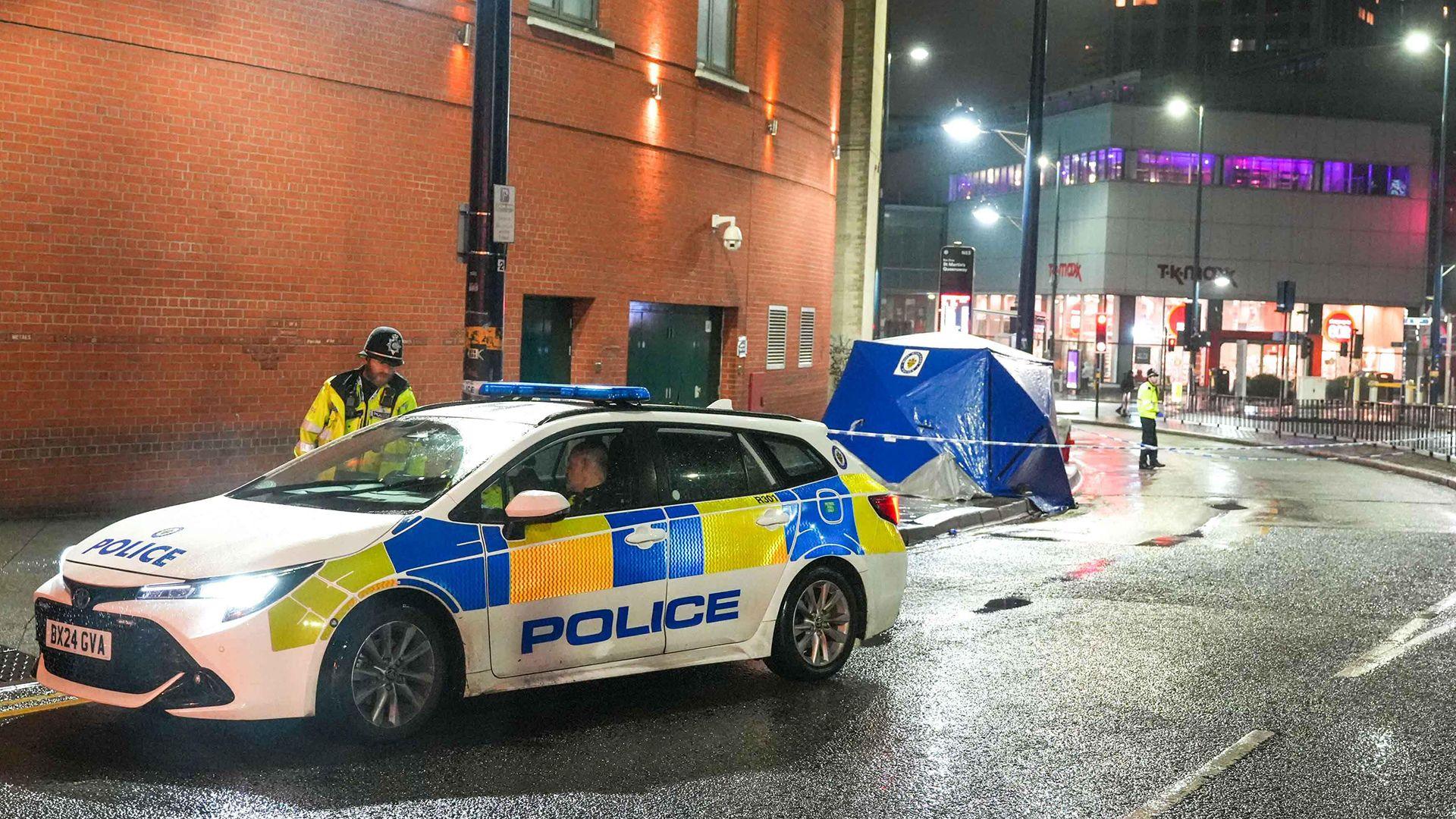 A police car is parked  in front of a blue forensics tent erected on the street outside Birmingham's Bullring shopping centre. It is at night and street lights are on.