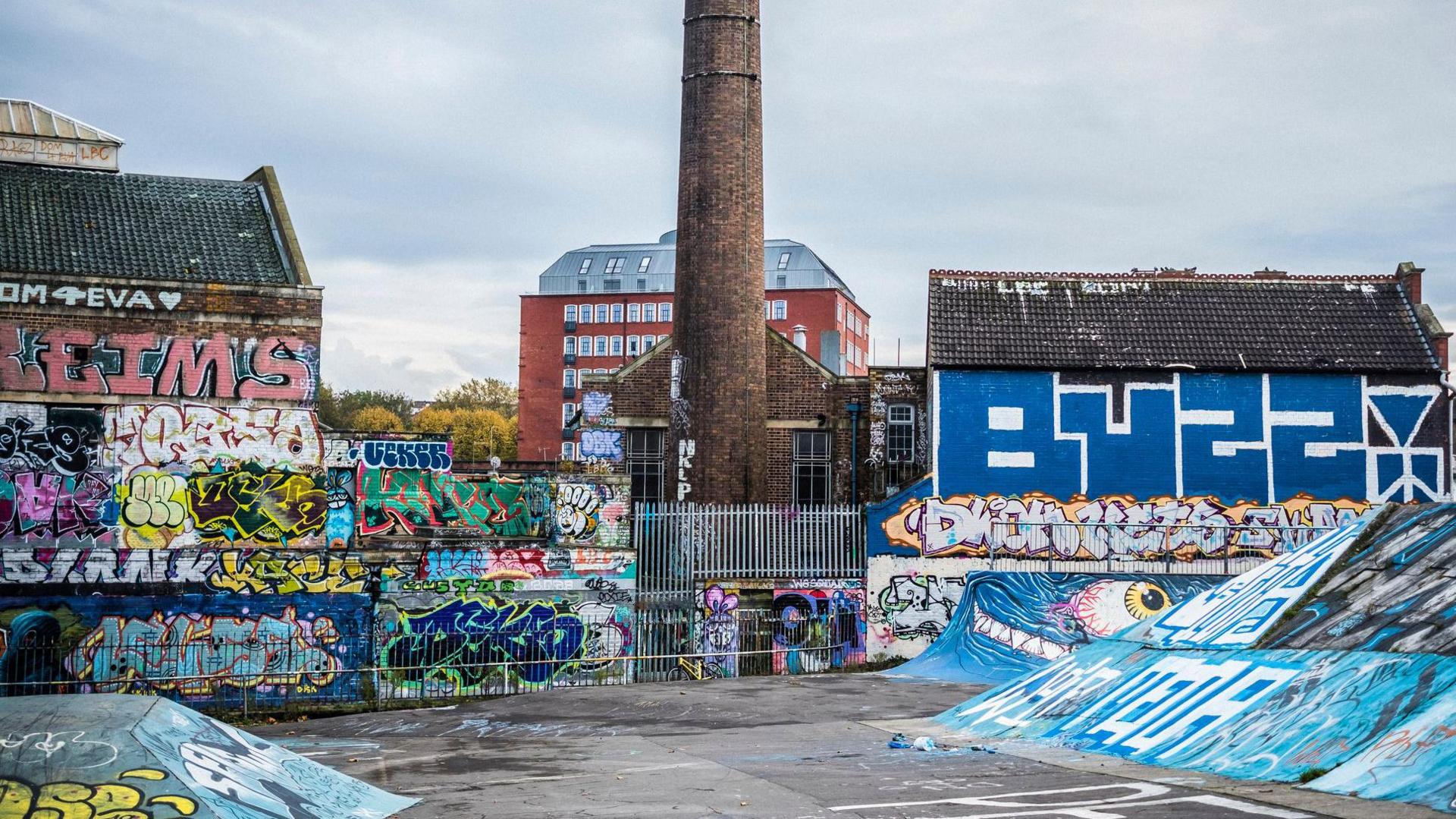 Dean Lane Skate Park covered in colourful graffiti. The only part of the park not covered too heavily in graffiti is the tarmac floor. Buildings and an old smoke chimney can be seen in the background.