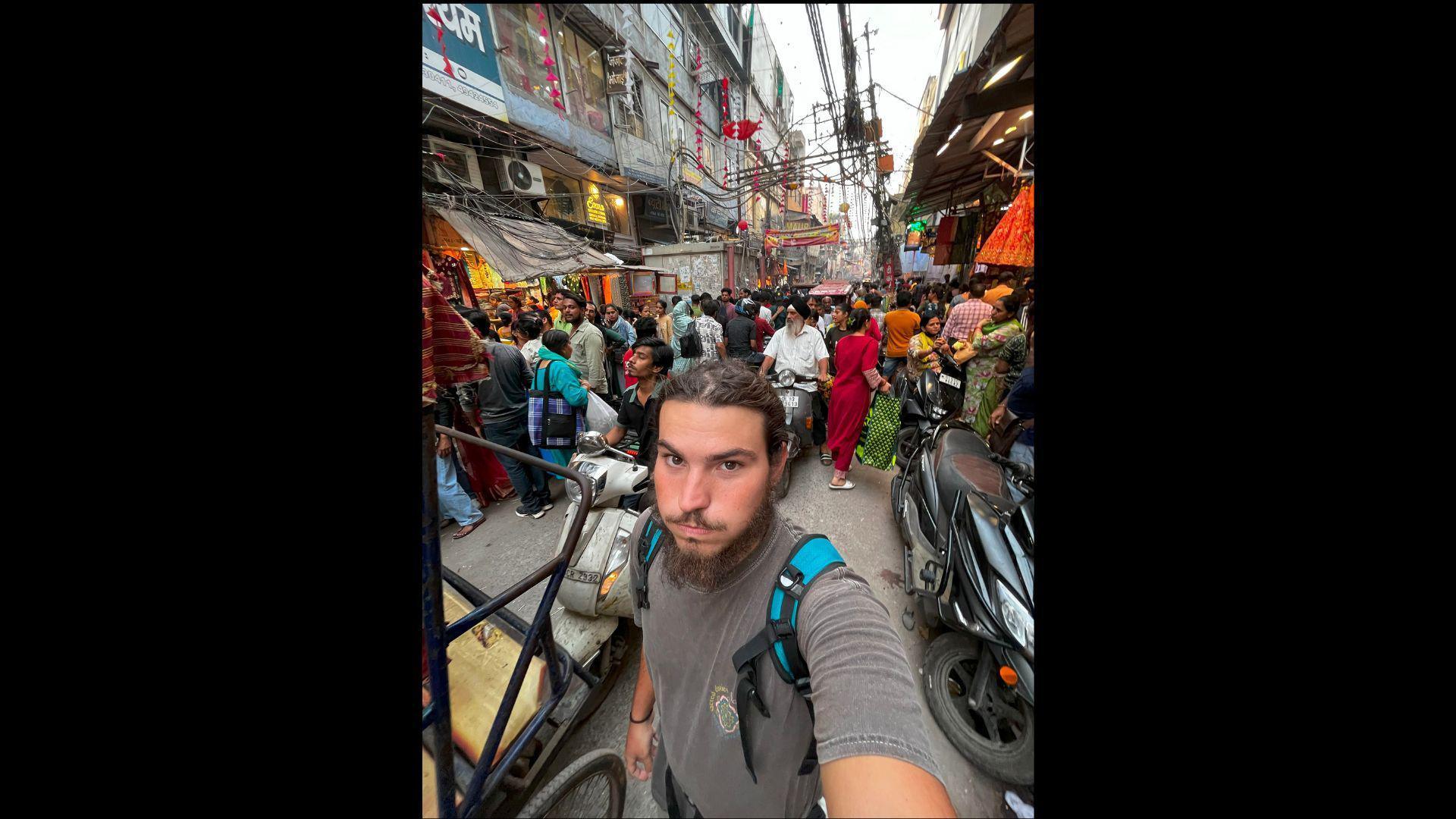 A man in a grey t-shirt, with a backpack, is standing in a street in New Delhi and celebrating the Diwali festival.