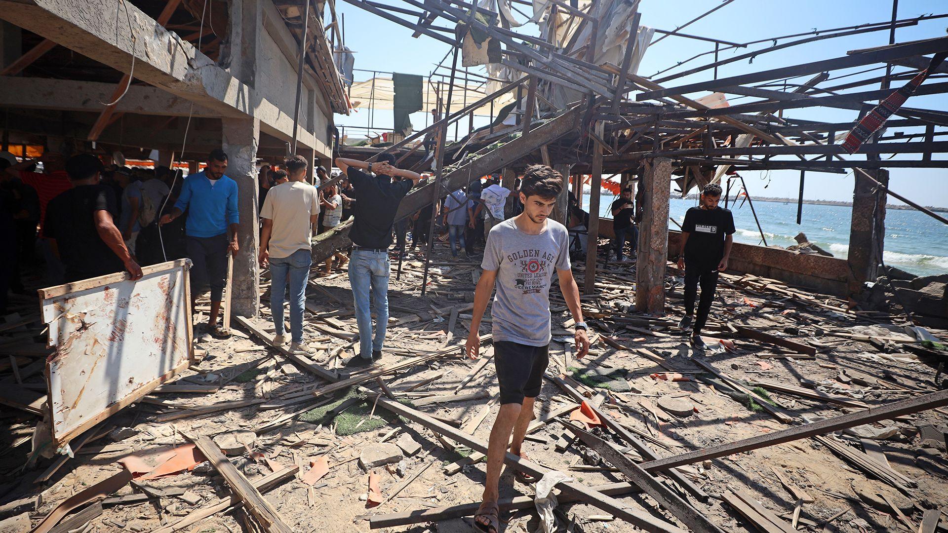 A view of the destruction after air strikes on a coffee shop in Gaza. People are pictured examining the debris with a young man walking out of the cafe in the foreground, and the sea and blue sky in the background, taken on 30 June.