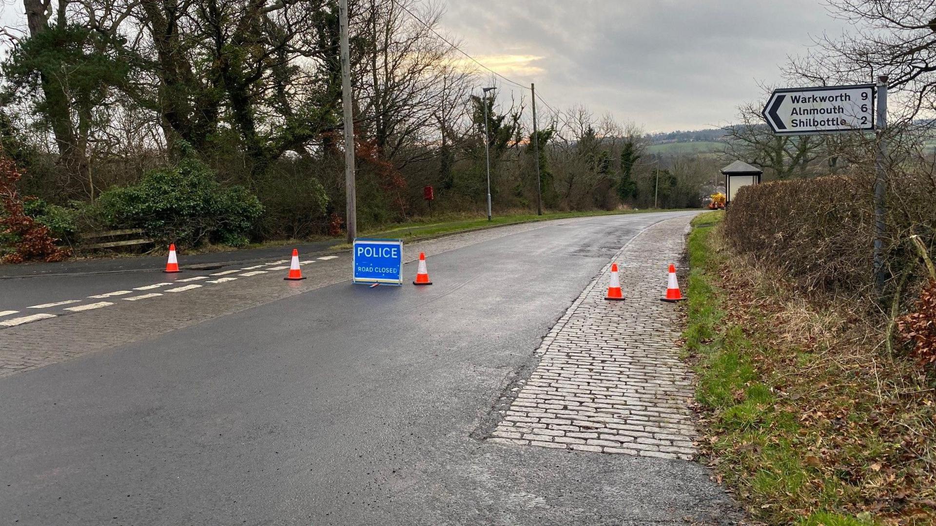 A rural road with cobbles on on side and a large blue sign from the police saying road closed 