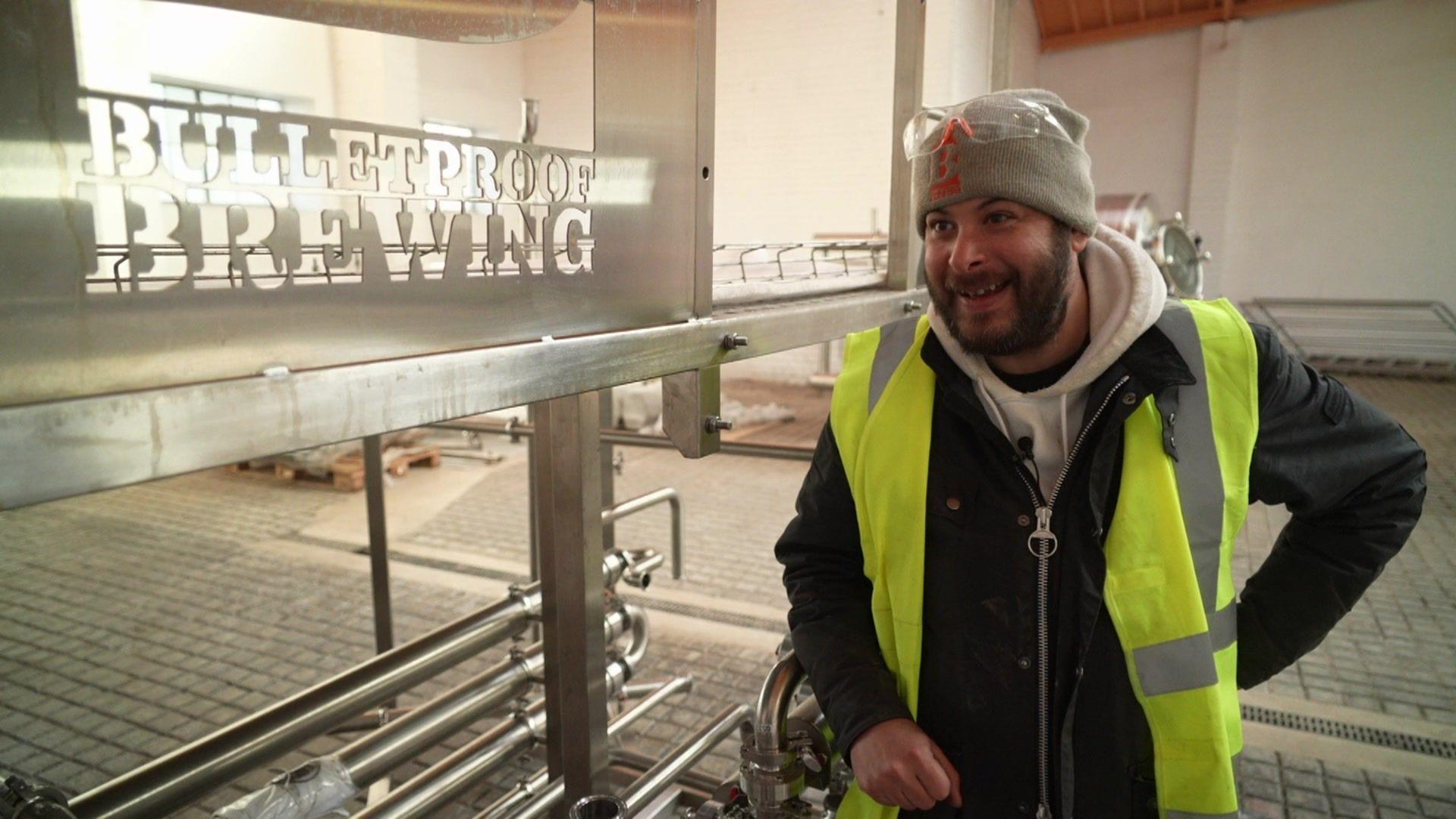 A man in a fluro vest and and a grey beanie next to a sign that says Bulletproof Brewery 