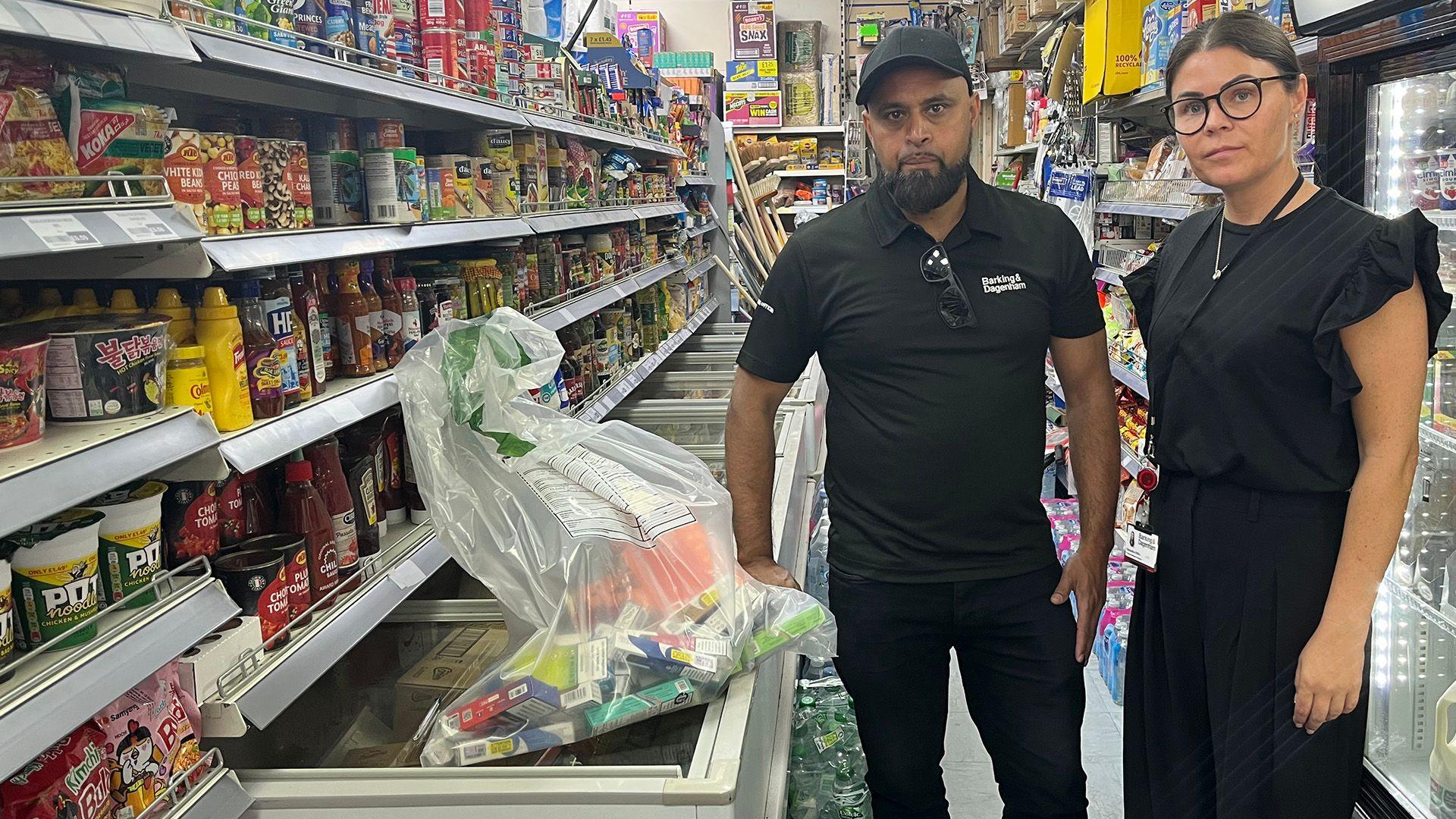 Nazir Ali and Samantha Veira stand in the aisle of a grocery store. Nazir is wearing a black polo shirt and dark pants, while Samantha is wearing a black dress. The aisle is filled with various food products on shelves, including canned goods, sauces, and snacks. A plastic bag filled with confiscated items sits atop a freezer in front of them.
