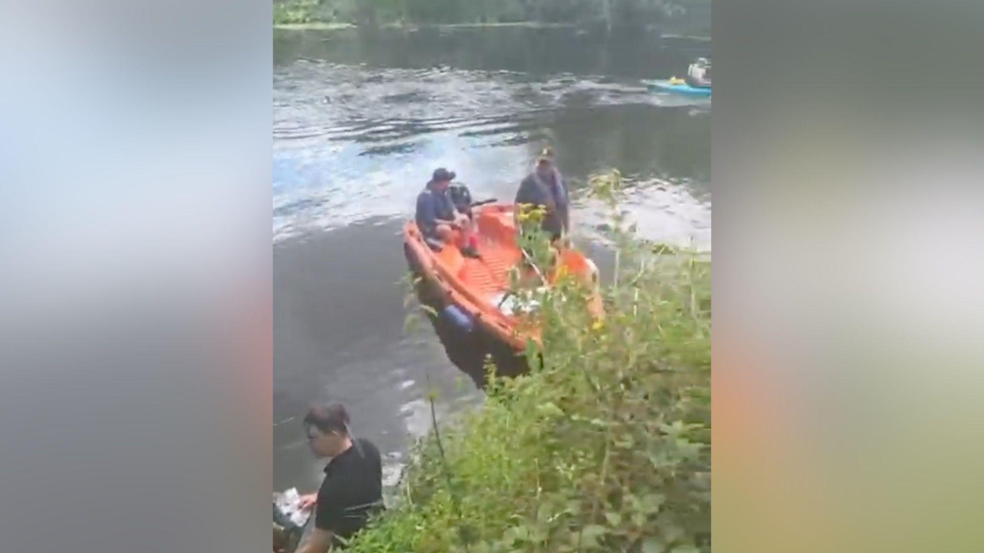 An orange rescue boat on the river Avon in Evesham. It has a man sitting in it, with another standing alongside. In the foreground is vegetation growing on the riverbank, while a man in a black T-shirt stands next to it in the water.