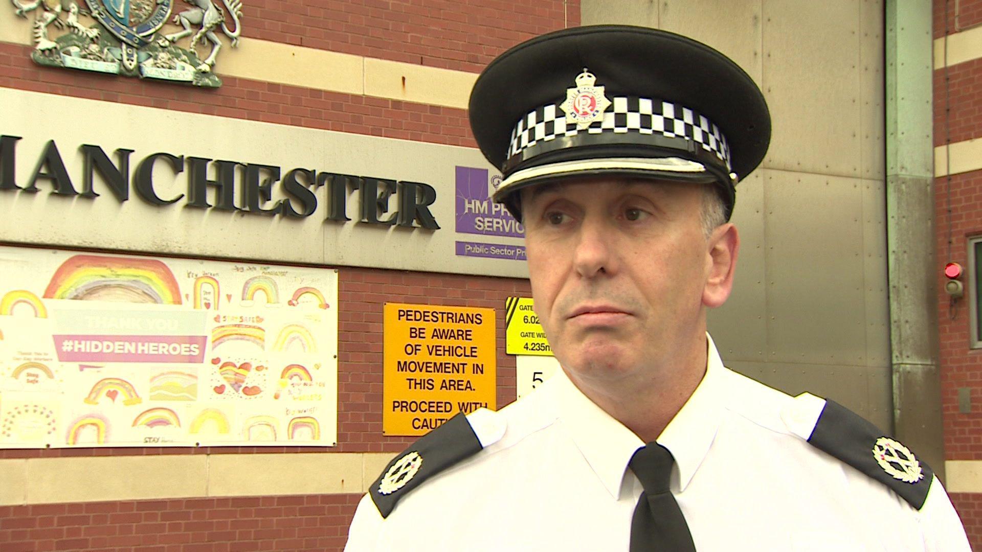 A GMP constable stands out a prison, wearing a police hat and shirt and black tie, looking off to the left of the camera. 