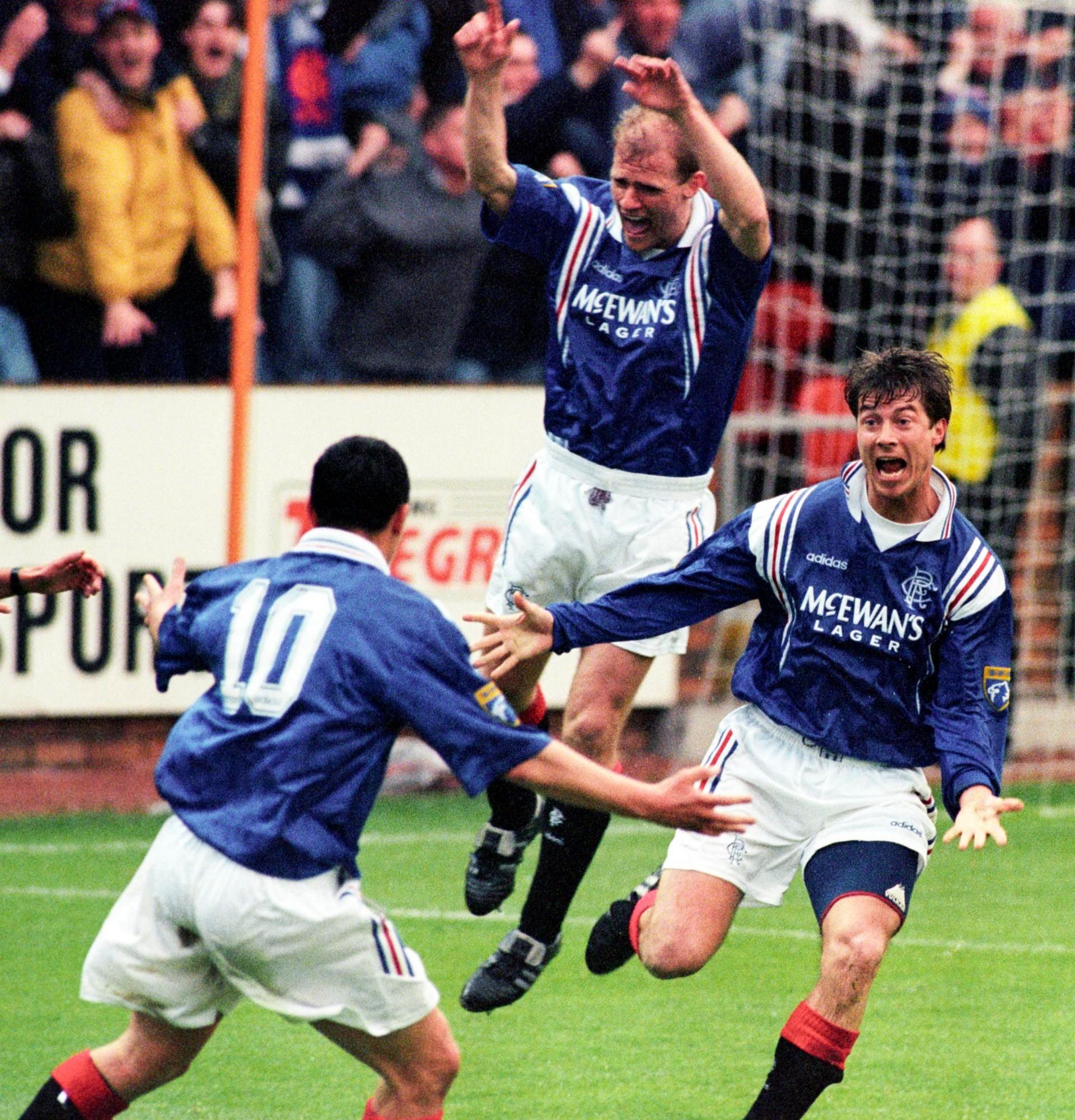 Rangers player Brian Laudrup with two teammates wearing a blue shirt and white shorts celebrating after scoring a goal. Laudrup is running towards the camera while his teammate in the background jumps in the air.
