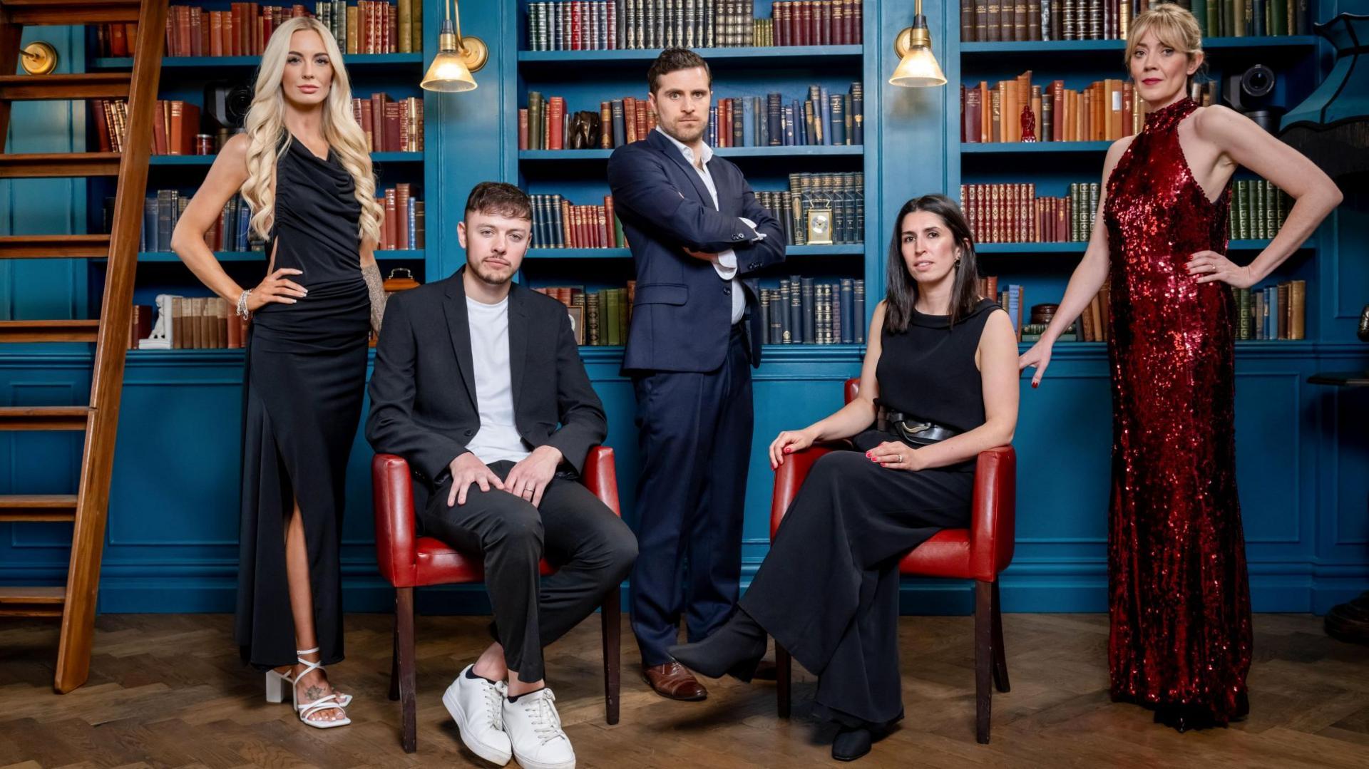 The five finalists of the Traitors in formal wear in a library with blue walls and a wooden floor 