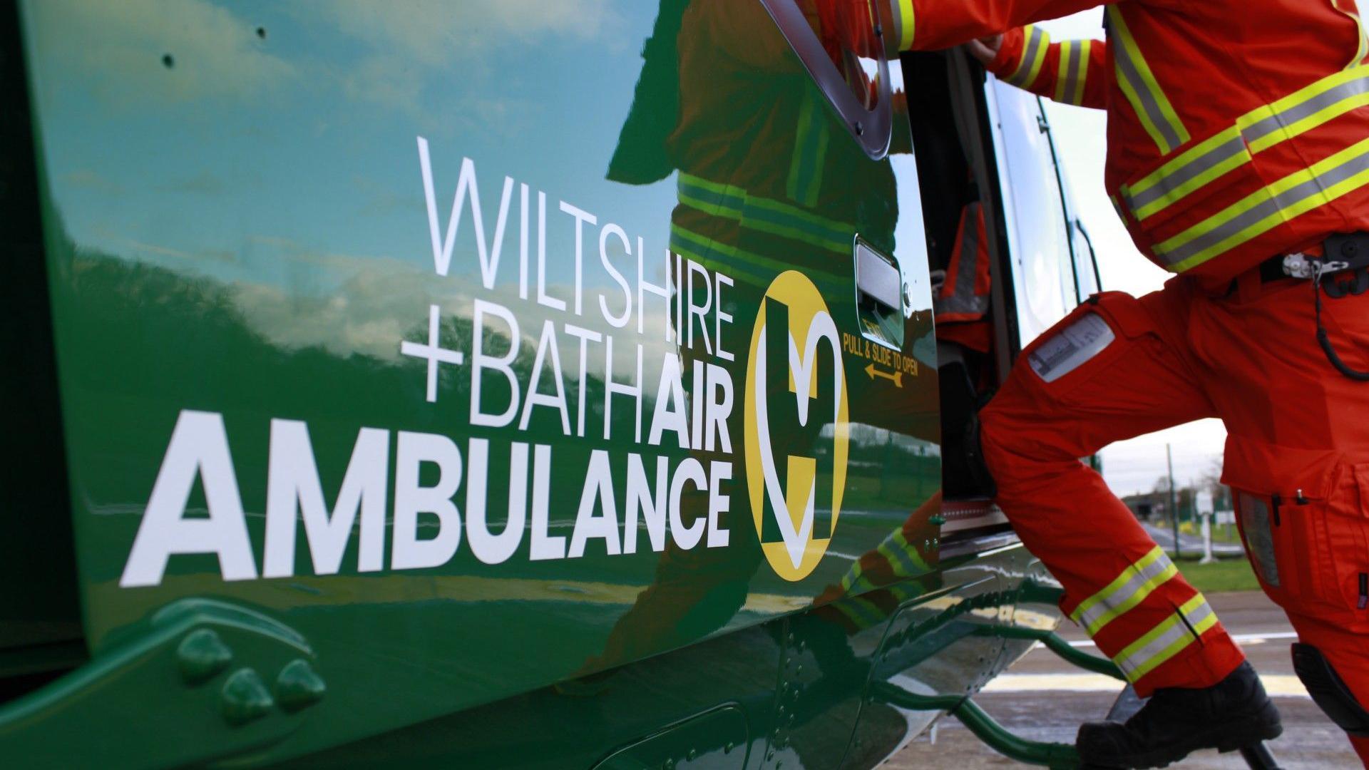 The Wiltshire and Bath Air Ambulance branding on the side of a helicopter and a emergency personnel stepping into it in uniform