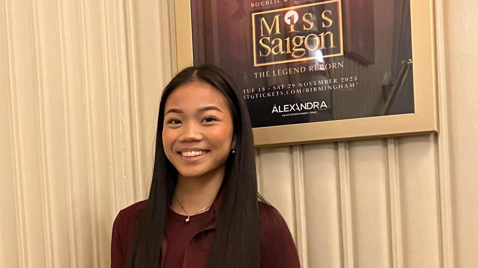 A girl with long straight black hair is pictured in a cream coloured room wearing a dark red T-shirt and dark red hoodie.