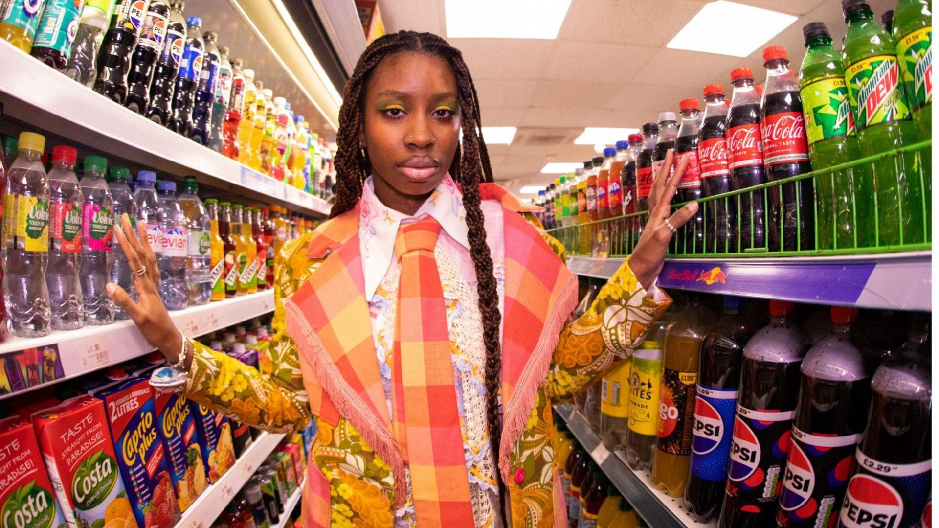 A woman standing between shop isles with long dark braids and wearing a multi-coloured jacket and tie