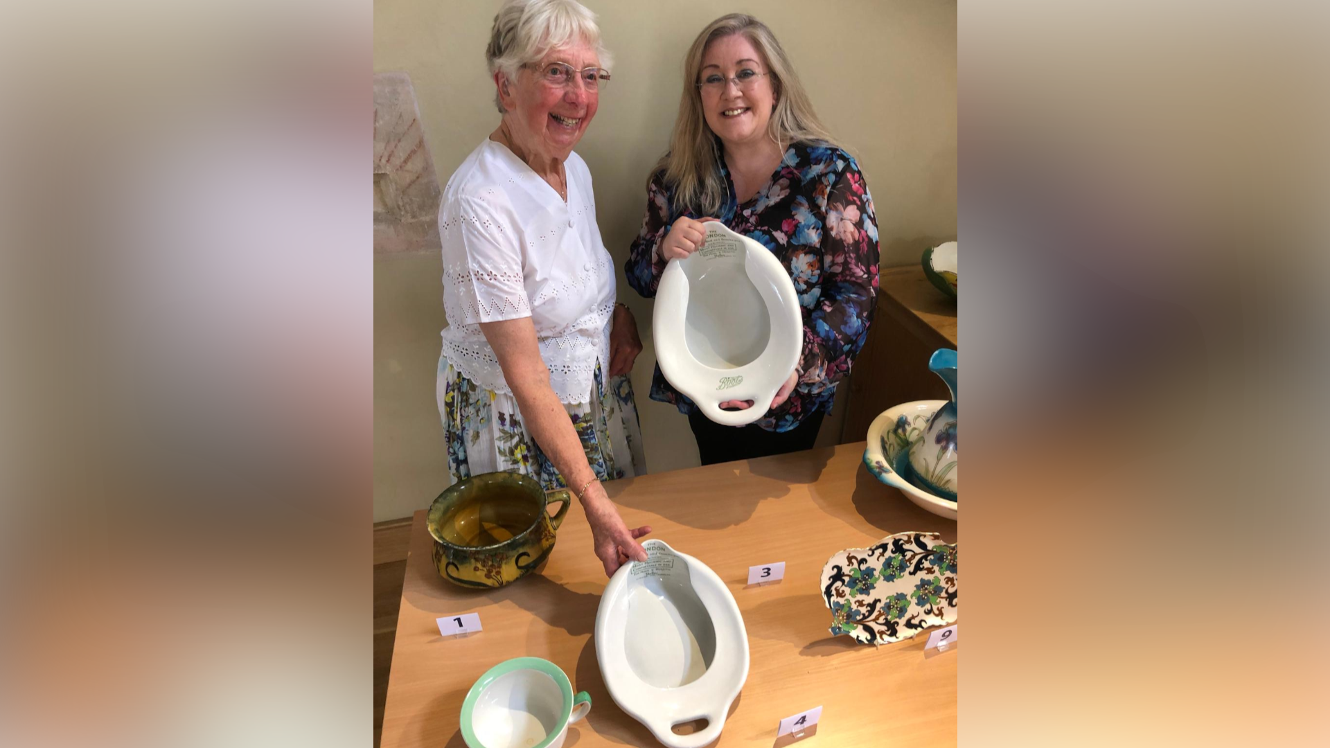 Mary Jacobs smiles while standing next to Julie Stevenson. Mary is looking away from the camera and is wearing a white blouse. She has short grey hair and is wearing glasses. Julie is smiling as she looks at the camera while holding a bedpan. She is wearing a floral blouse and has long blonde hair and glasses.