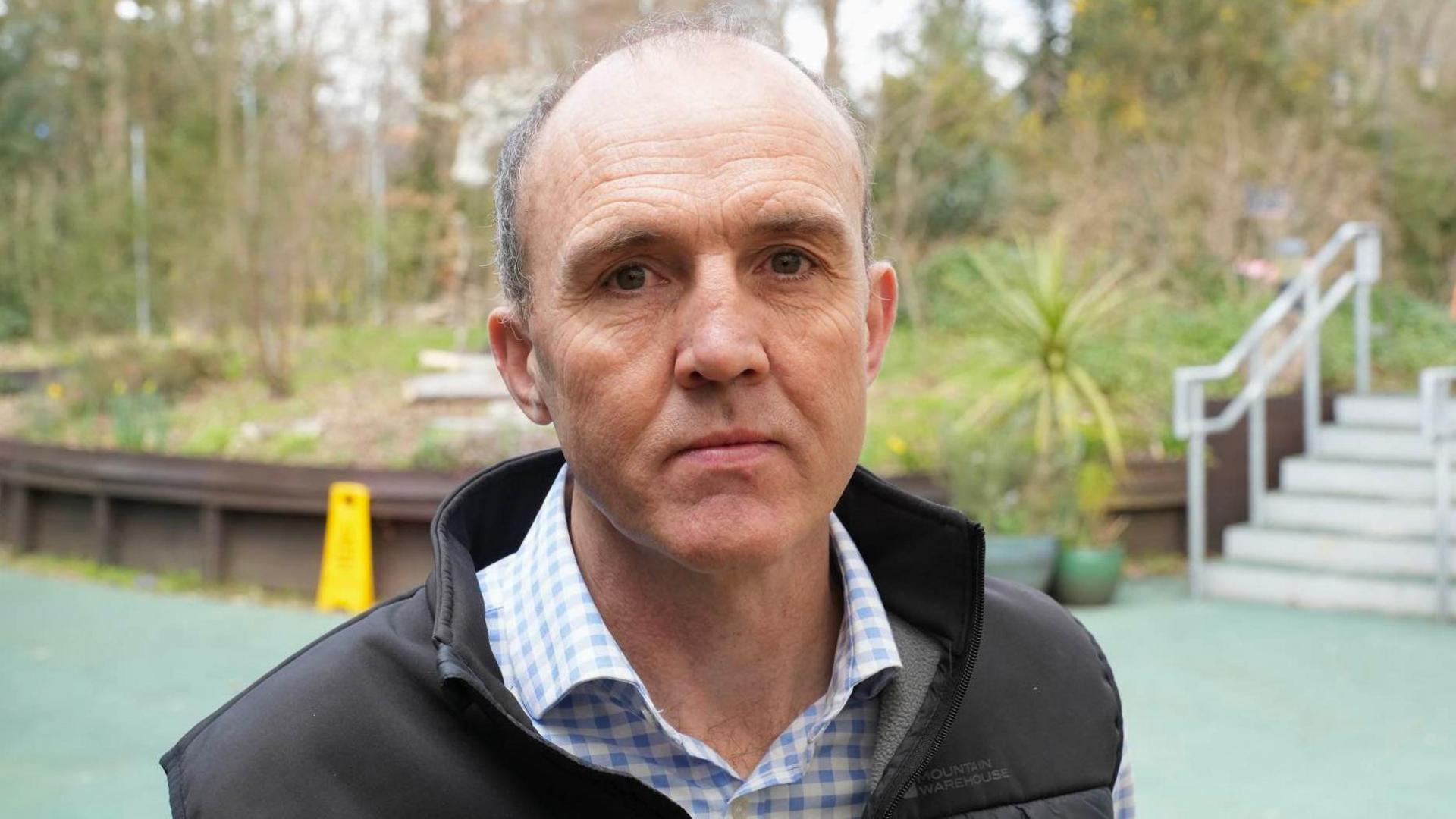 Ian Nicolson wearing a blue checked shirt and a black gilet. He has short grey hair and is cleanly shaven. He is standing in front of a garden.