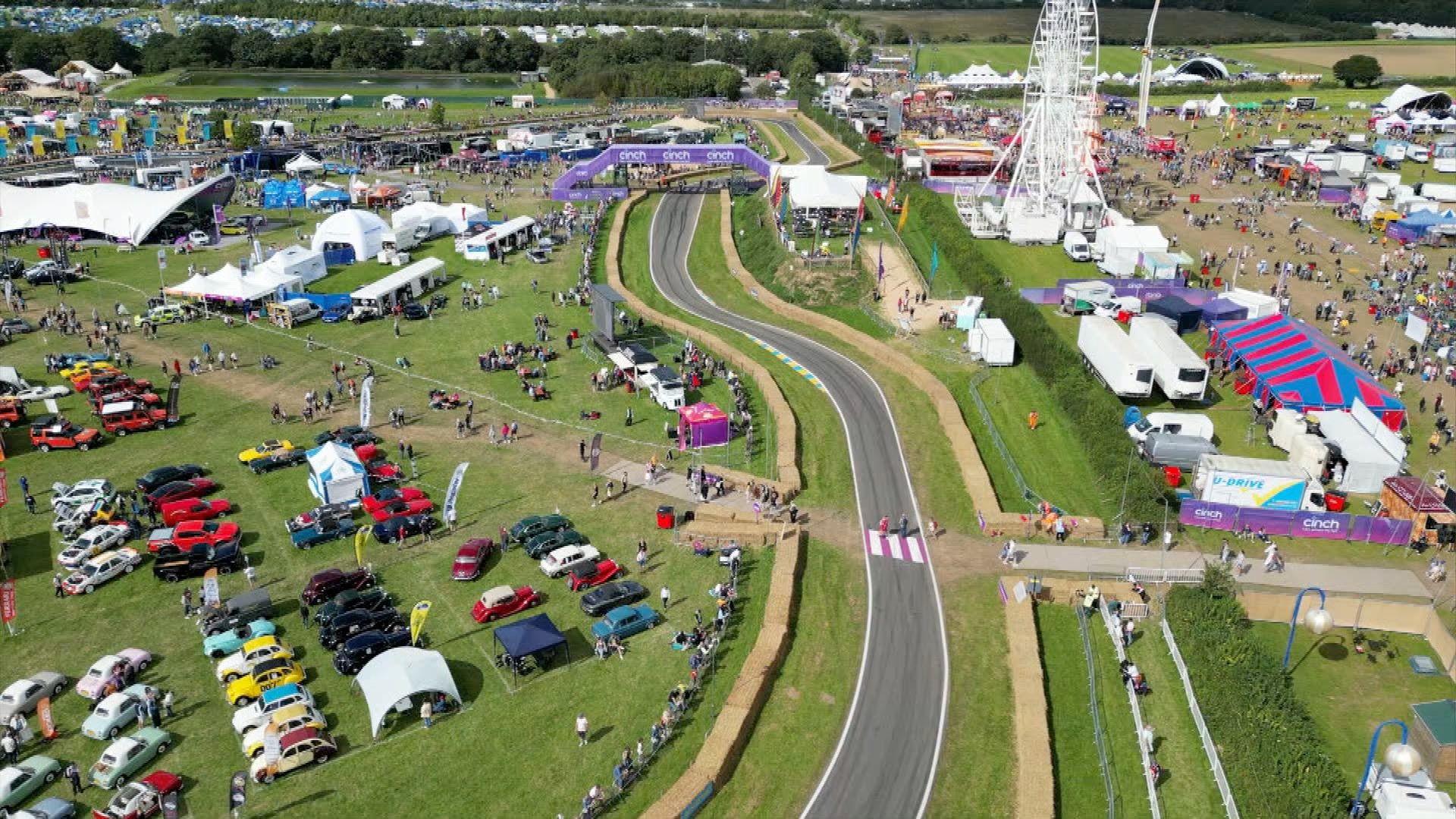 An aerial view of the festival in green fields, with parked cars on the left, a racing track running through the middle and a large white ferris wheel behind and to the right,