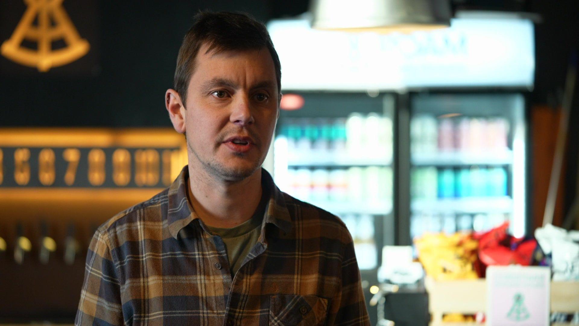 A man wearing a brown and blue stripy shirt talking off camera. Behind him is beer taps and a drinks fridge.