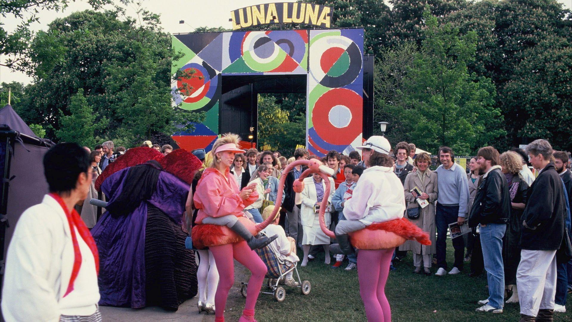 Two performers dressed as flamingos touch beaks at the center of a crowd.