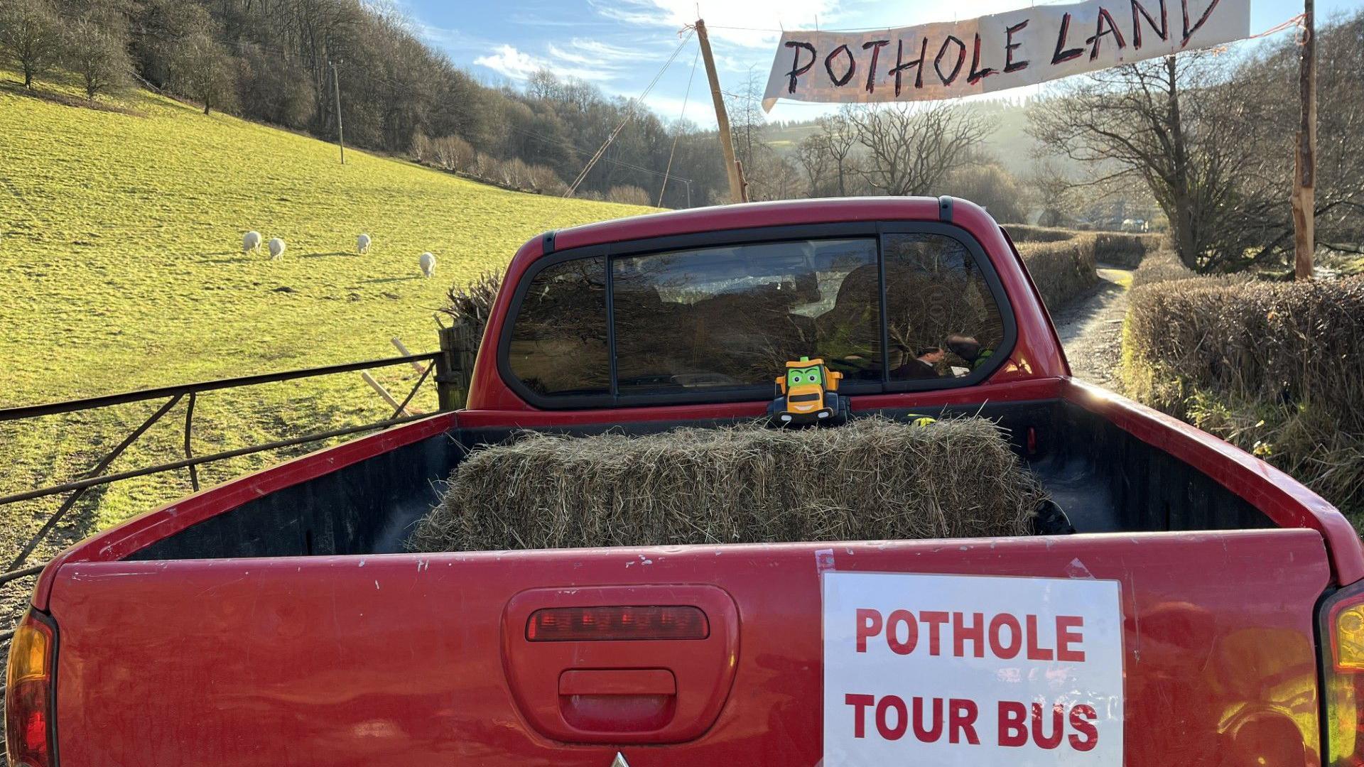 A red pick up truck from the back, pointing up a lane over which hangs a banner which says Pothole Land.  The truck has a sign on the back which says POTHOLE TOUR BUS.  There is a hay bale and an orange toy tractor in the back of the pick-up. There are sheep in the sunny field at the side of the lane. 