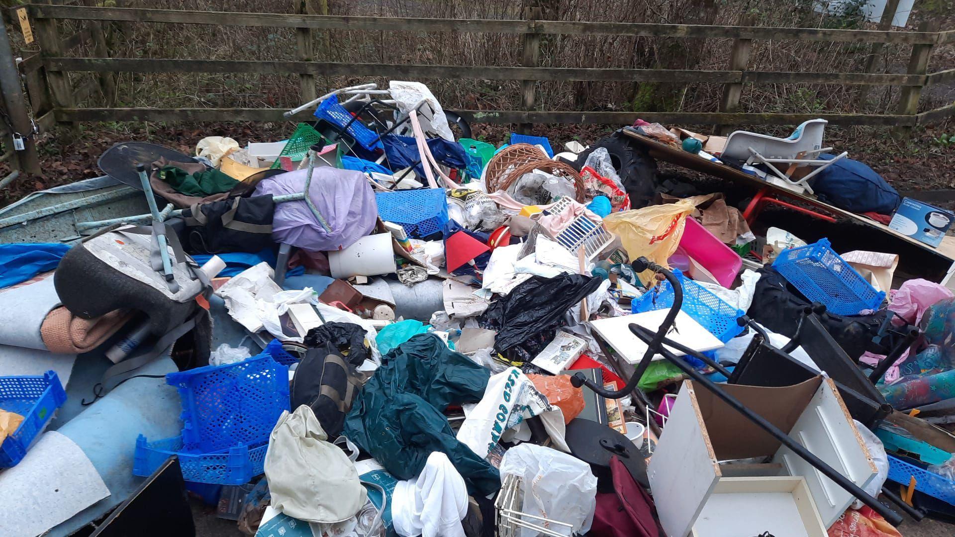 A close up of the fly-tipped rubbish, including large metal bars, several bags of different material and an office chair. A wooden fence can be seen in the background. 