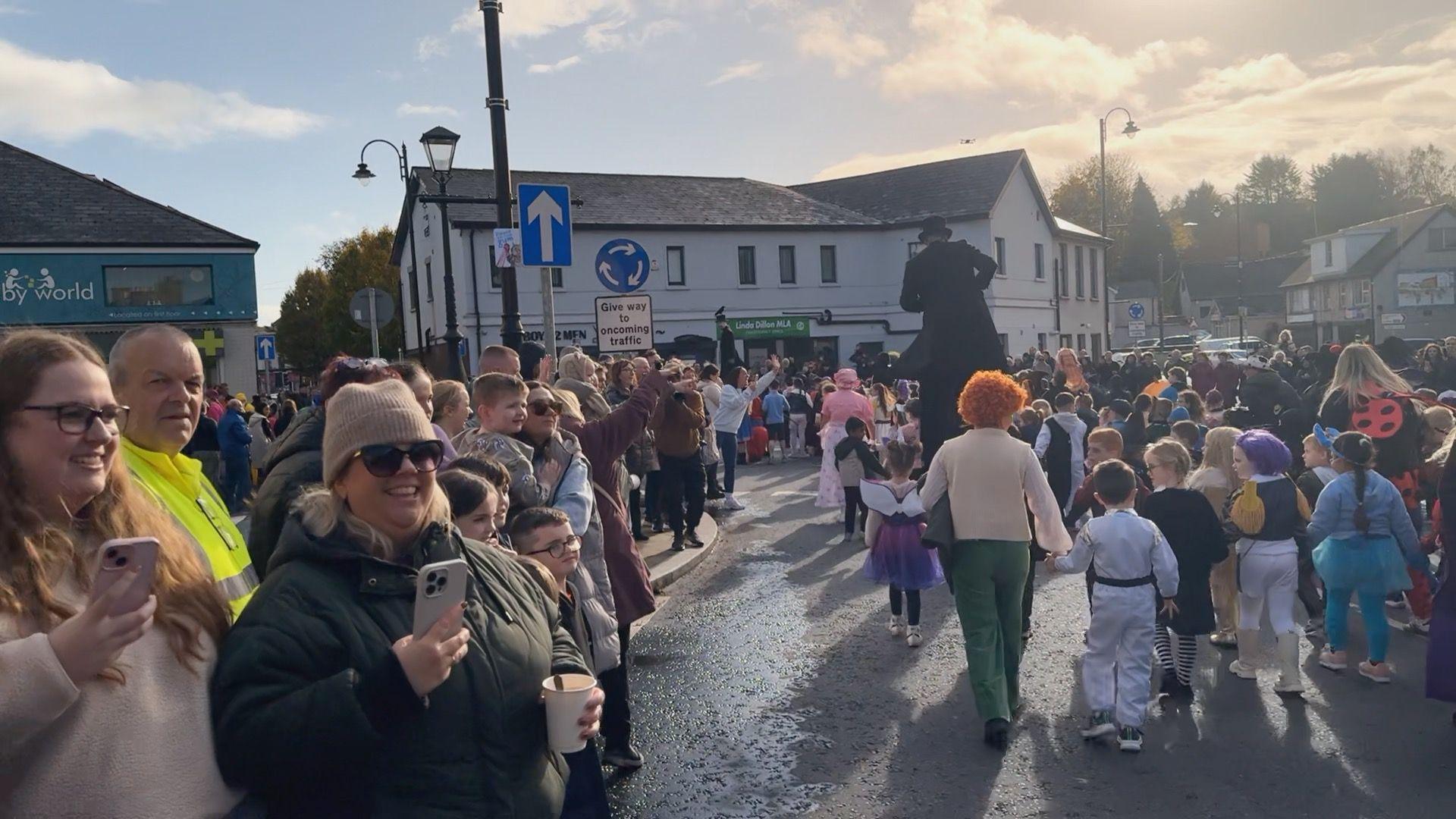 A wide shot shows the parade passing through the town, on the left of the screen are adults watching the parade, some holding mobile phones to take photos.