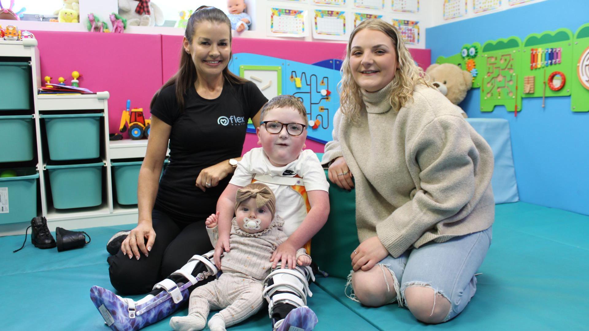 Two women smile as they kneel on either side of two children - a five-year-old boy and a baby girl - in a children's play area with soft furnishings in blue and pink, toys and storage shelves. To the left is therapist Lisa Speight, a woman with dark-brown hair, tied back, wears a black top with the word "flex" in blue letters, and black trousers. To the right, a woman with long blond curly hair wears a baggy cream jumper and light-blue ripped jeans.