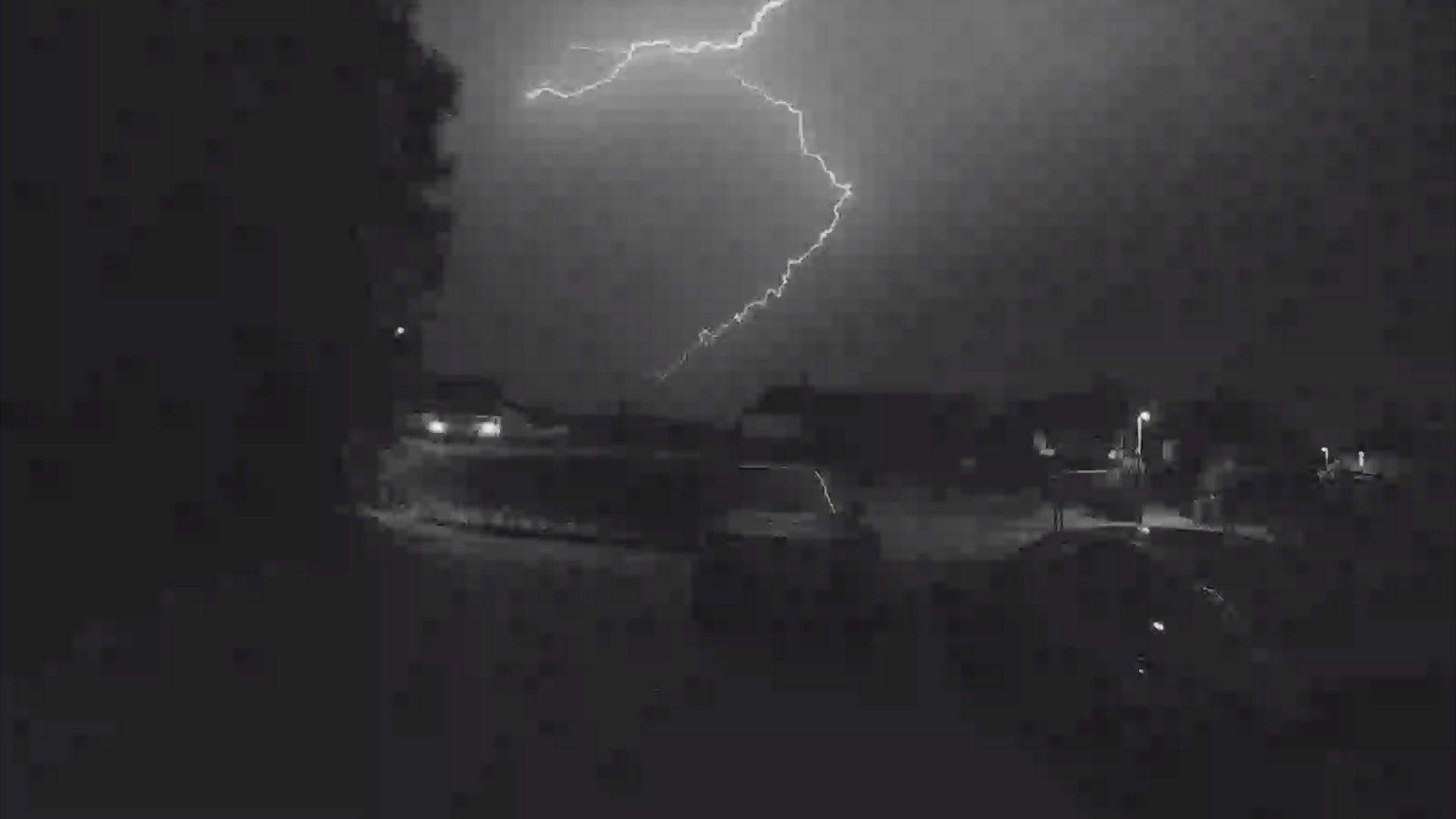 A black and white image, possibly taken from a doorbell camera, captures a large lightning bolt in the shape of a backwards C striking. It is a very dark image but a large hedge and a few cars can just about be made out in the foreground, with the rooftops visible in the background.