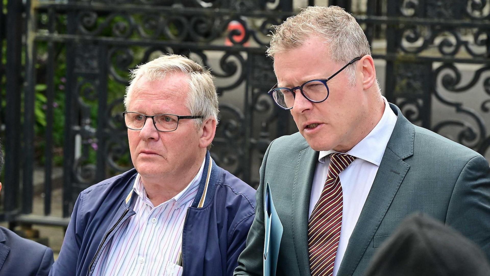 Three people stand behind two different microphones. The man on the right is grey-haired with blue glasses. The man wears a grey suit, white shirt and Brown striped tie. To his right is a grey haired man in black glasses, wearing a blue jacket and a blue and white striped shirt. To the men's right is a man in a navy suit and white shirt, has short brown hair and glasses. T