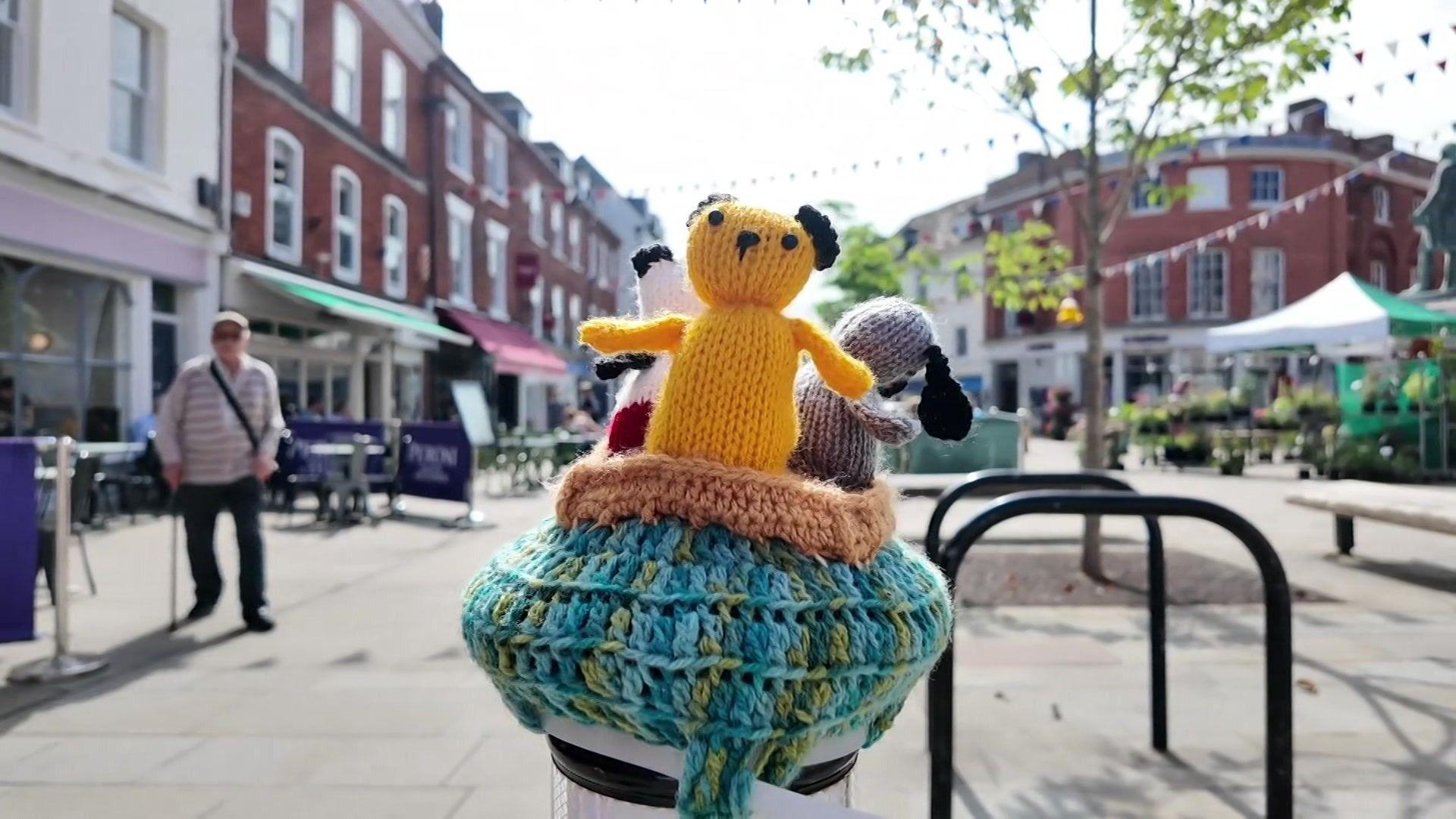 A woollen Sooty character on top of a bollard on a pedestrianised area with shops in the background.