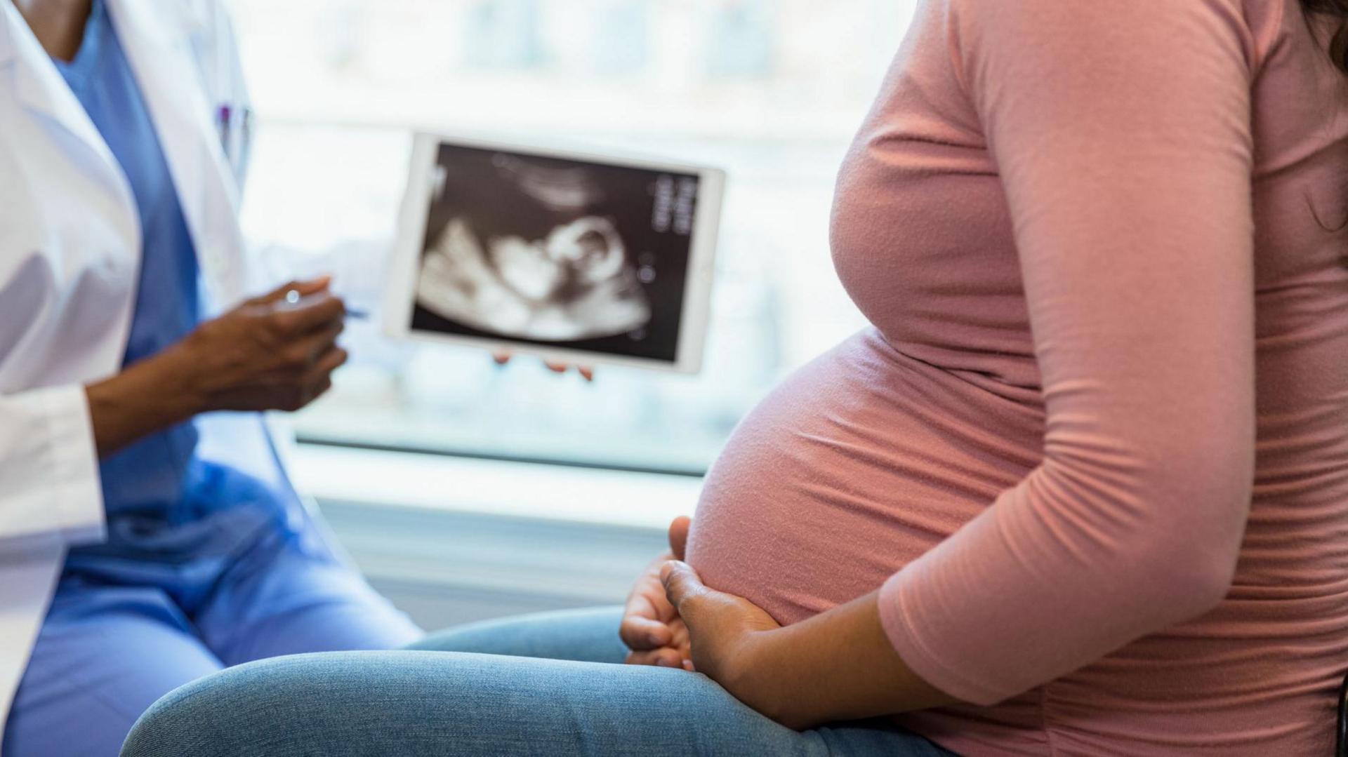 A heavily pregnant woman wearing blue jeans and a pink top sits side-on, talking to a healthcare professional who is showing her a baby scan on a tablet.