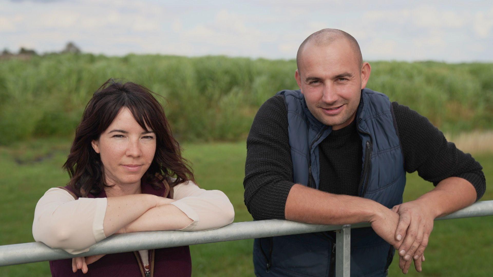 A man wearing a black t-shirt and blue gilet stands next to a woman wearing a white t-shirt and red gilet. They are leaning over a metal fence in a green field, with their arms resting on the fence. 