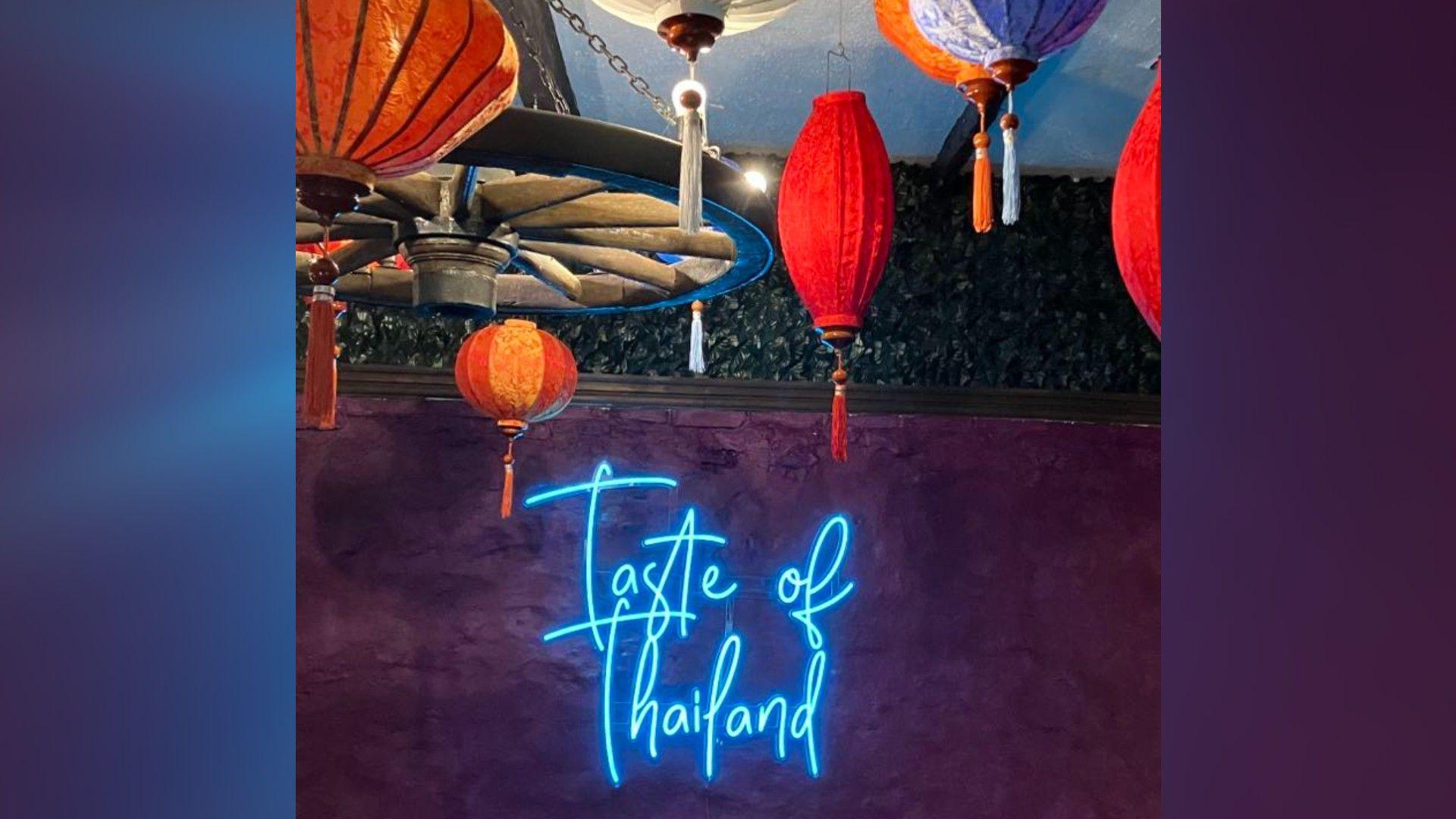 A purple wall with a blue-lit Taste of Thailand sign and colourful lanterns on the ceiling 