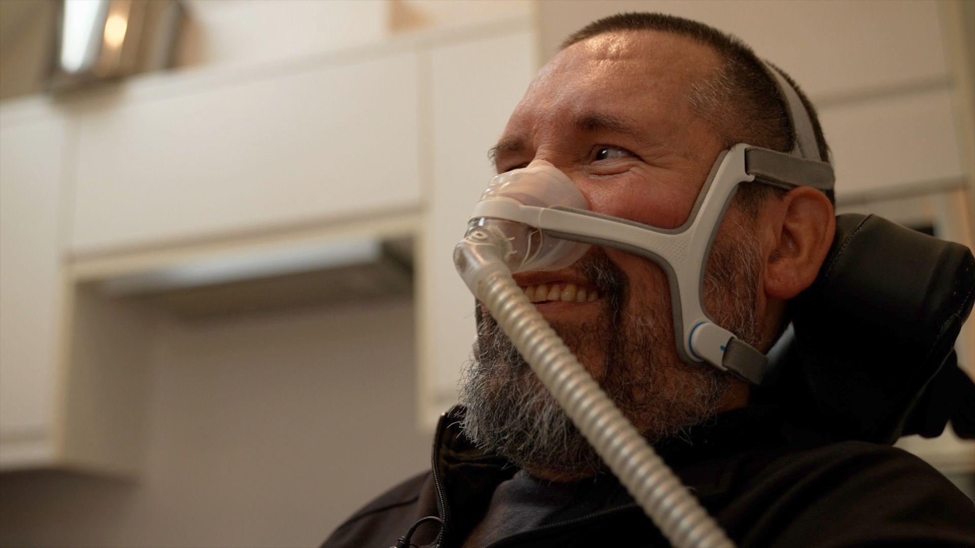 A smiling Nik Ward looks towards his family who are standing to the left, out of shot. He has a beard and is sitting in a wheelchair with a breathing mask across his face.